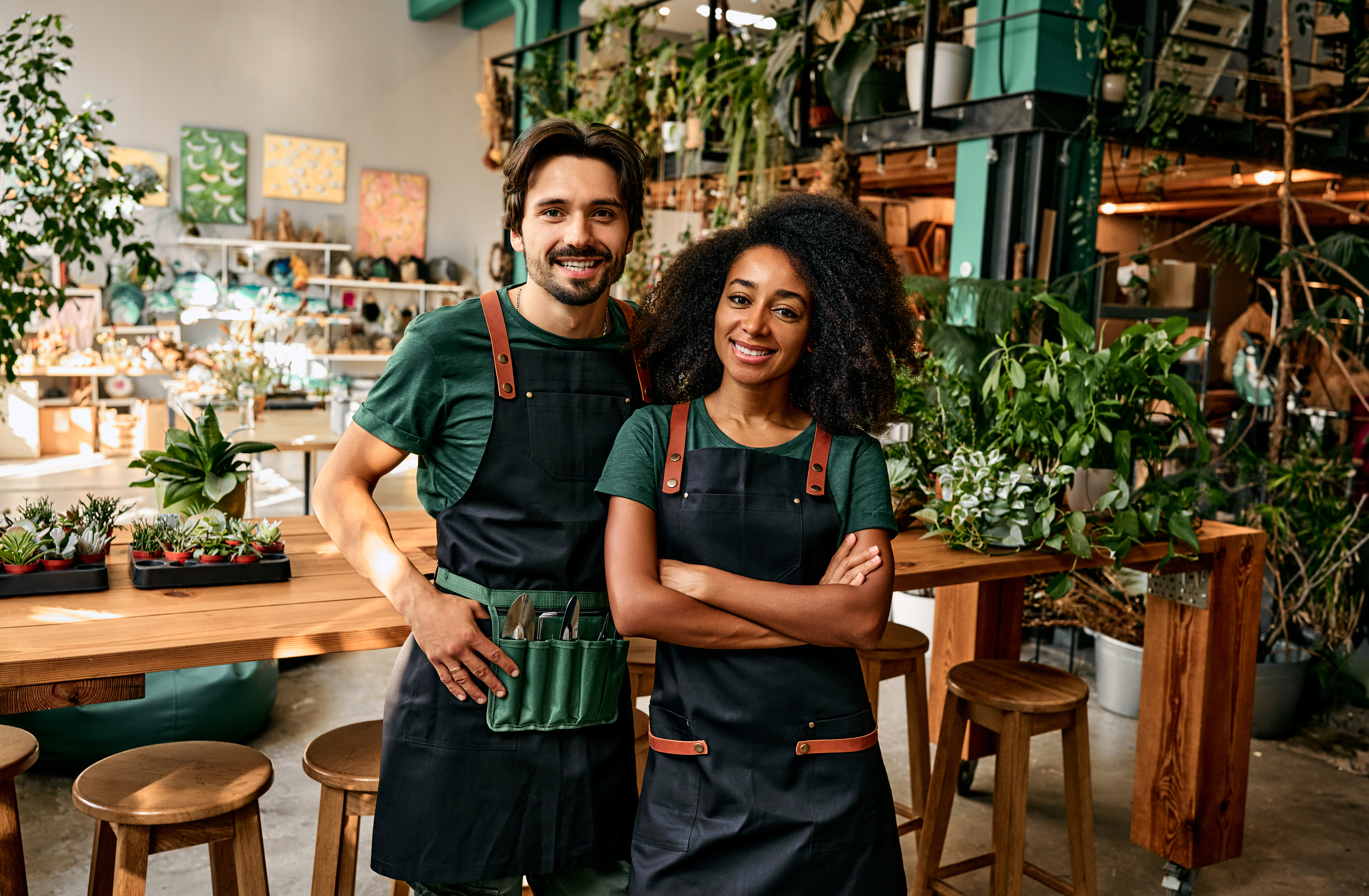 Small flower business. Front view of caucasian man and african american woman in aprons smiling at camera white standing at own shop. Small flower business. Front view of caucasian man and african american woman in aprons smiling at camera white standing at own shop.