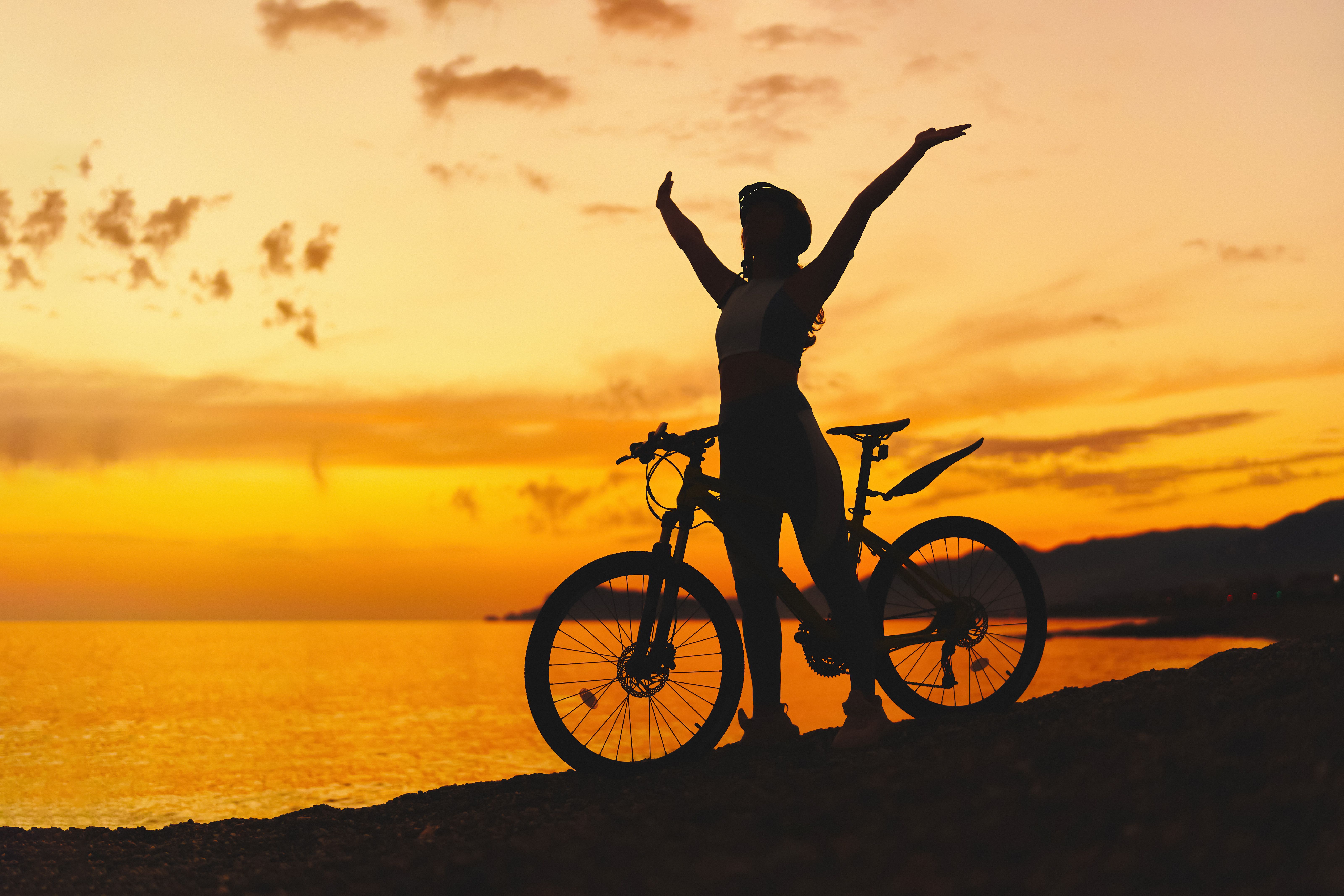 Silhouette of active one free happy joyful carefree inspired woman bicycler with open arms standing alone with bicycle outdoors against sunset sky
