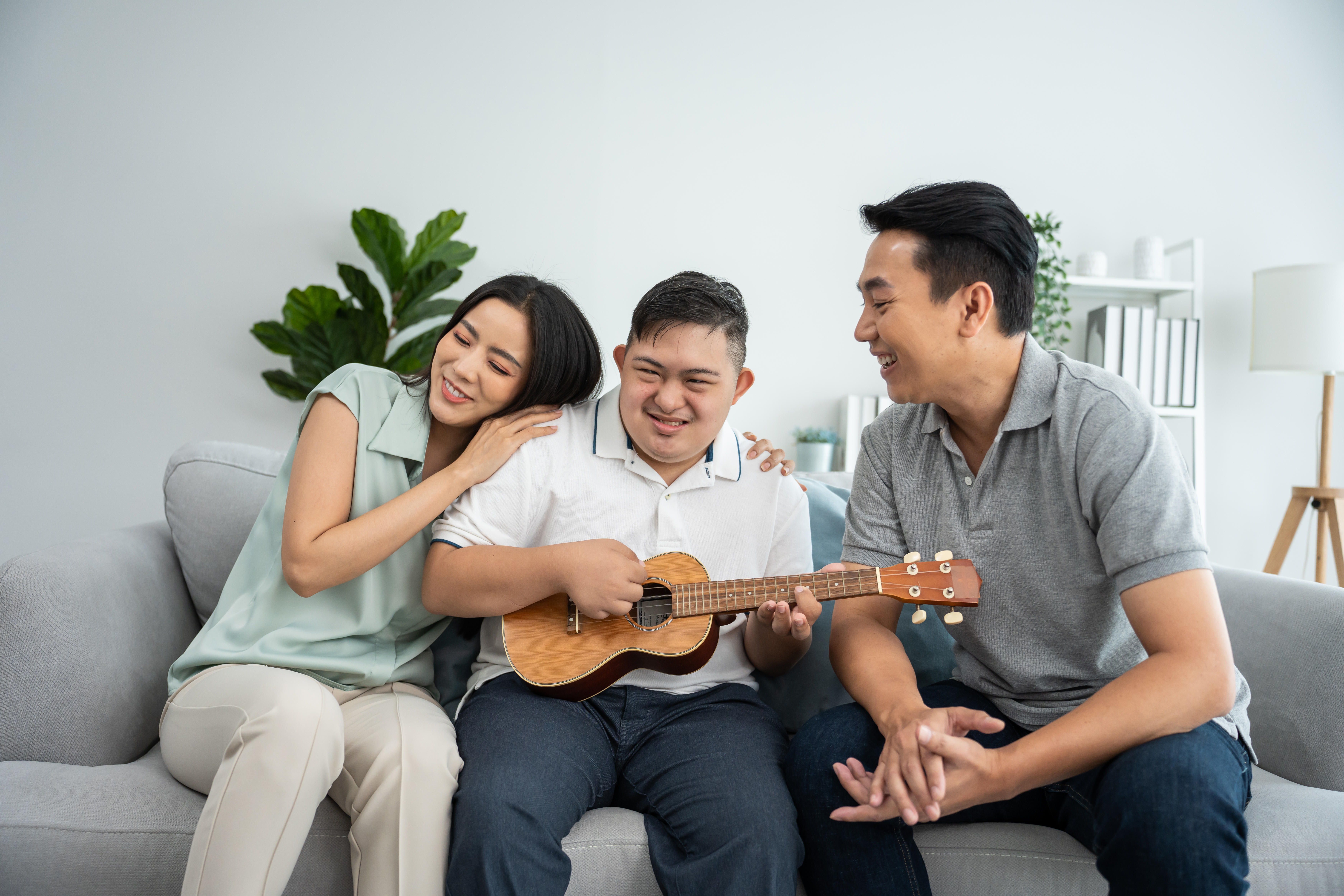 Asian attractive family playing with down syndrome son in living room. Beautiful loving mother and father take care and teaching young man with autism or special needs play ukulele indoors at home.
