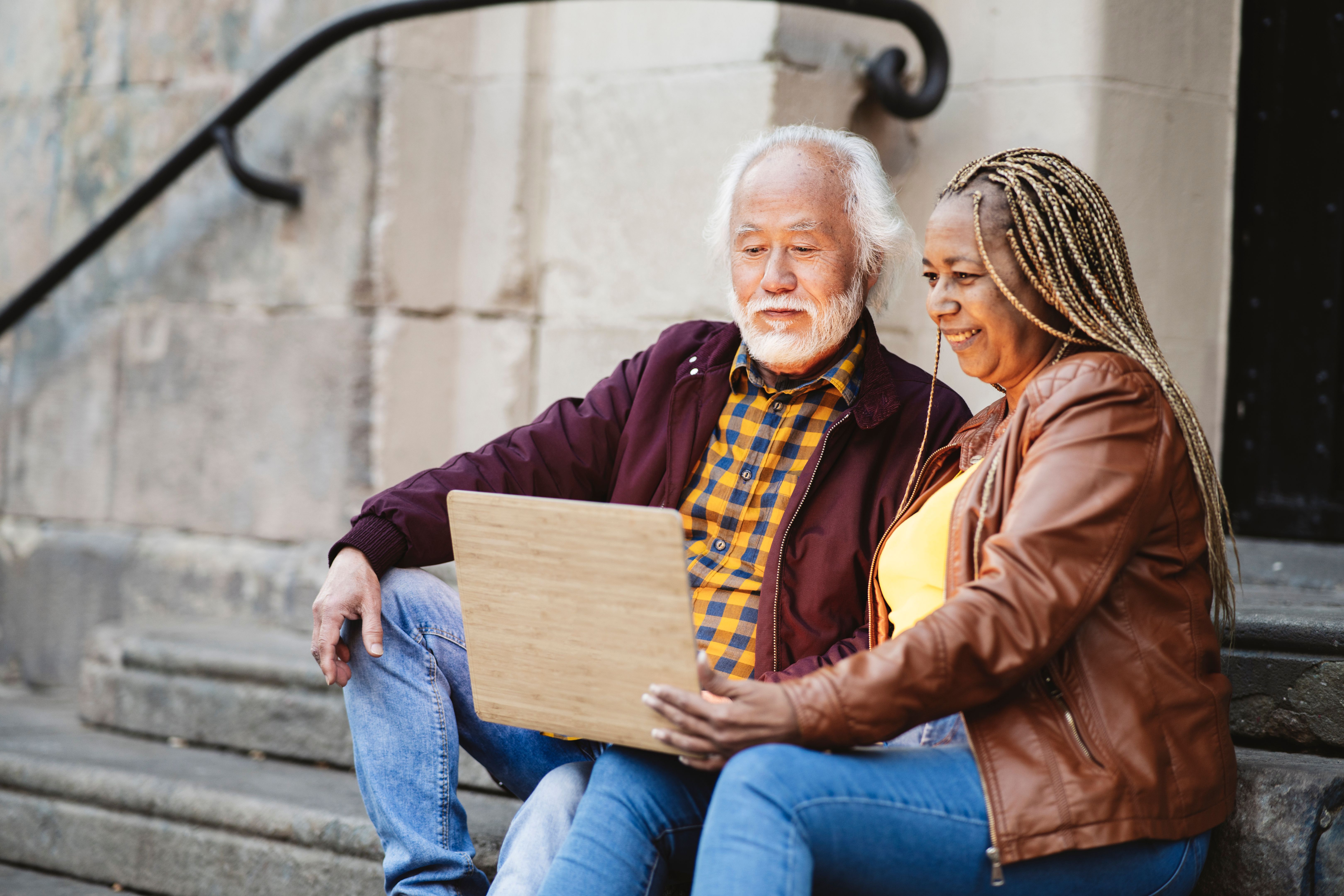Multiracial senior couple sitting on stairs looking at laptop in the city. Focus on asian man