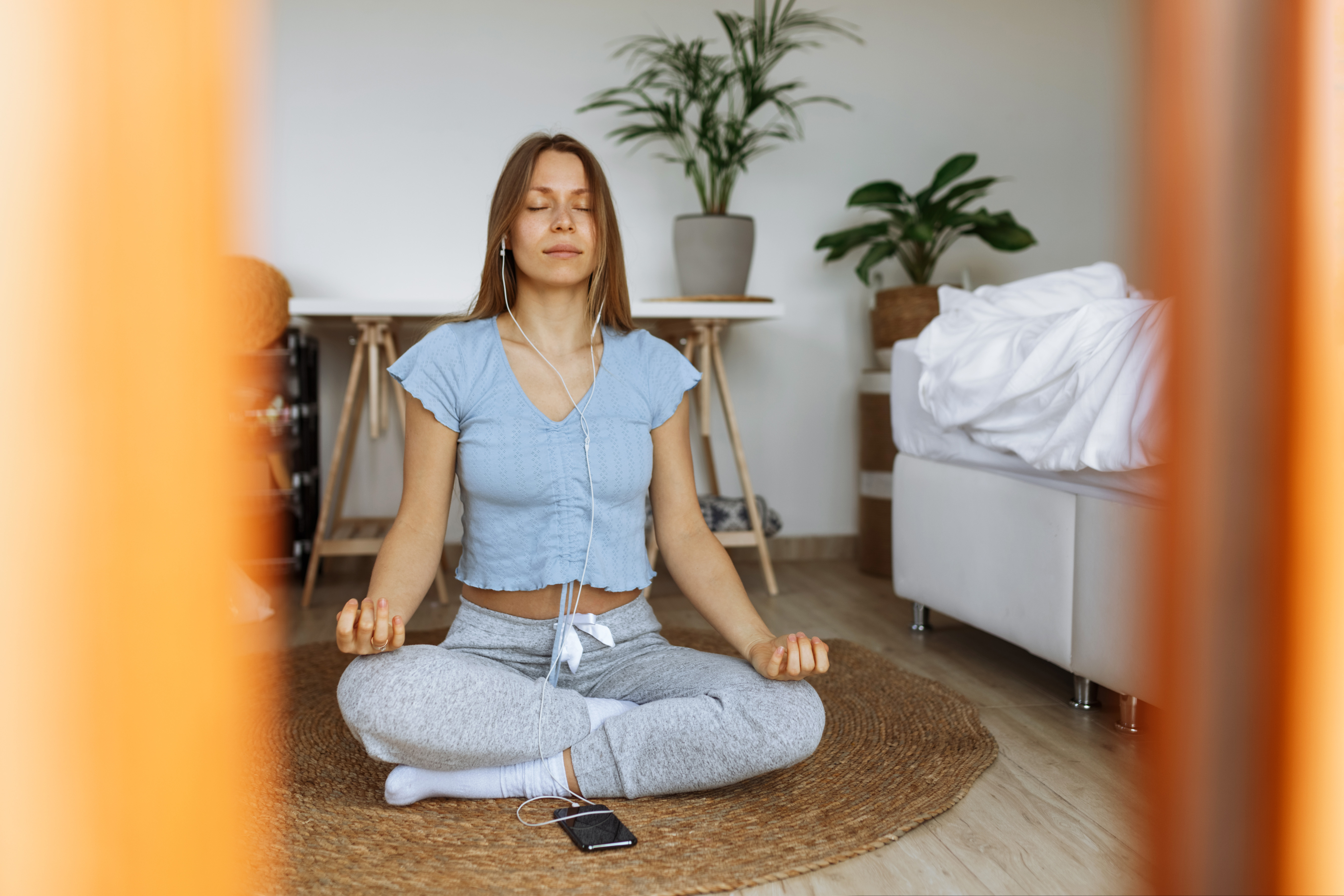 Trendy young woman wearing earphones meditating online and listening music on smartphone player app while sitting on floor at home,meditation app as part of mindfulness morning routine,peaceful mood