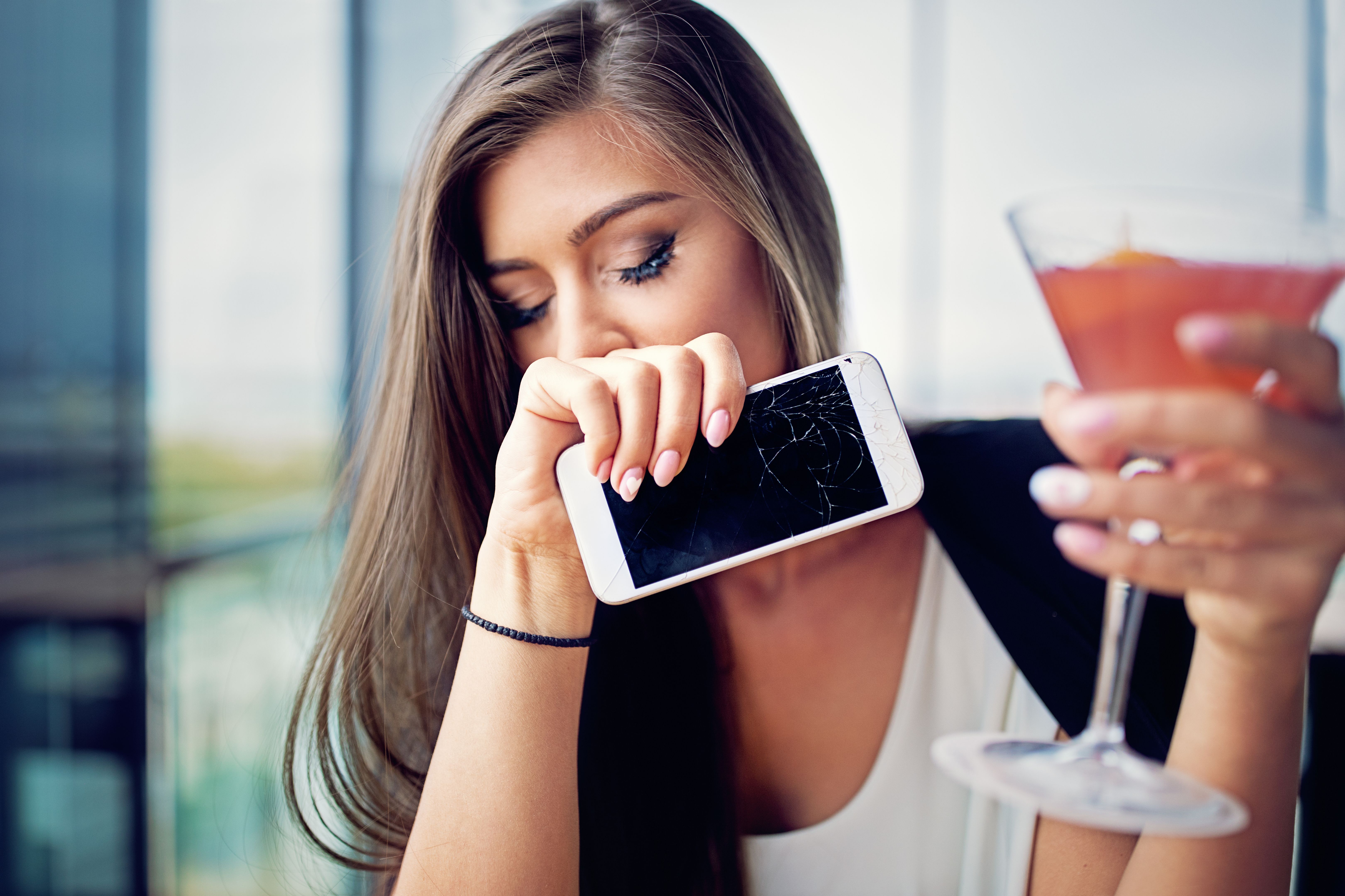 Desperate woman is drinking cocktail and holding her broken phone in the bar