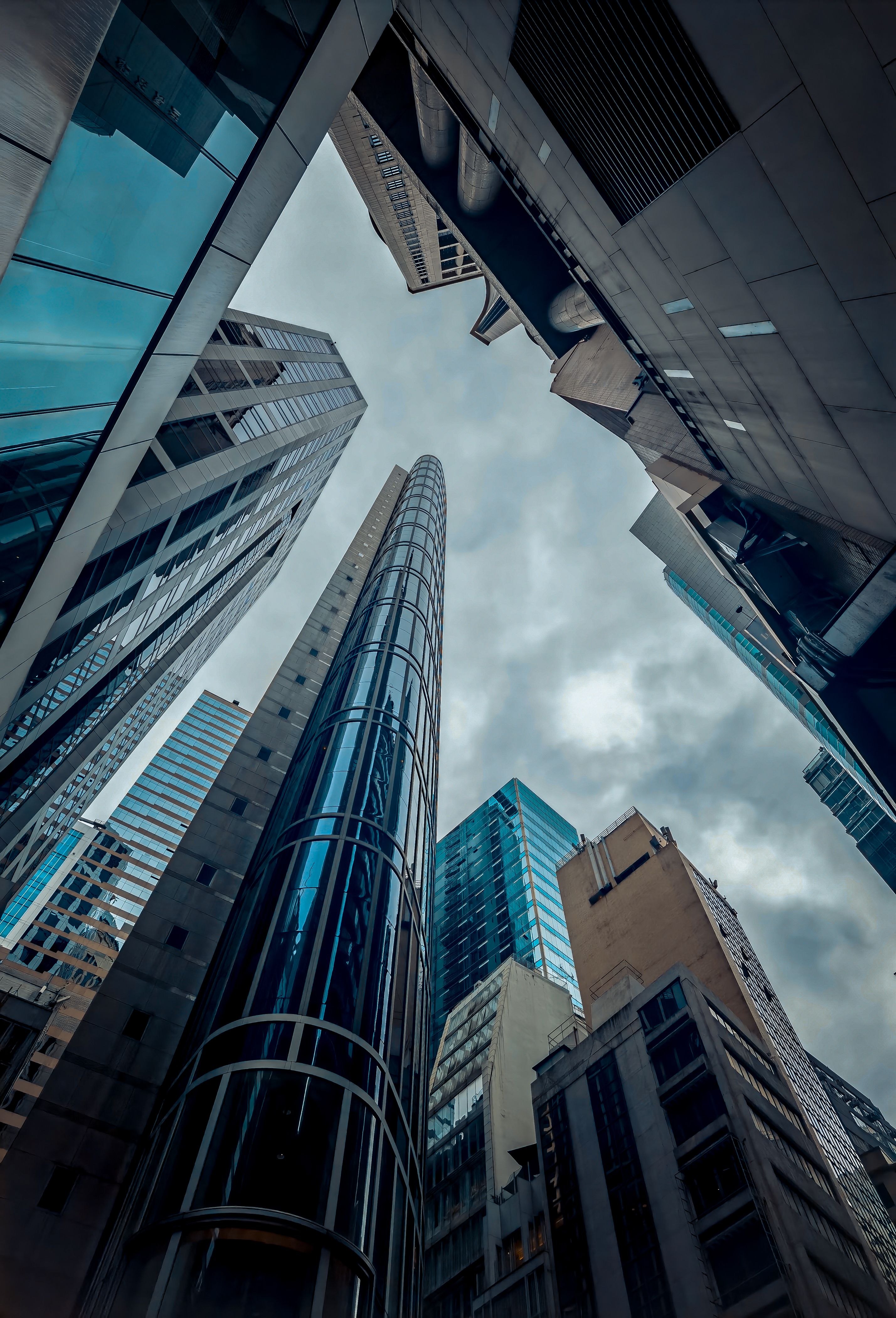 A low-angle, close-up view of towering skyscrapers in Hong Kong, piercing a cloudy sky.  The image emphasizes the city's dense, vertical architecture.