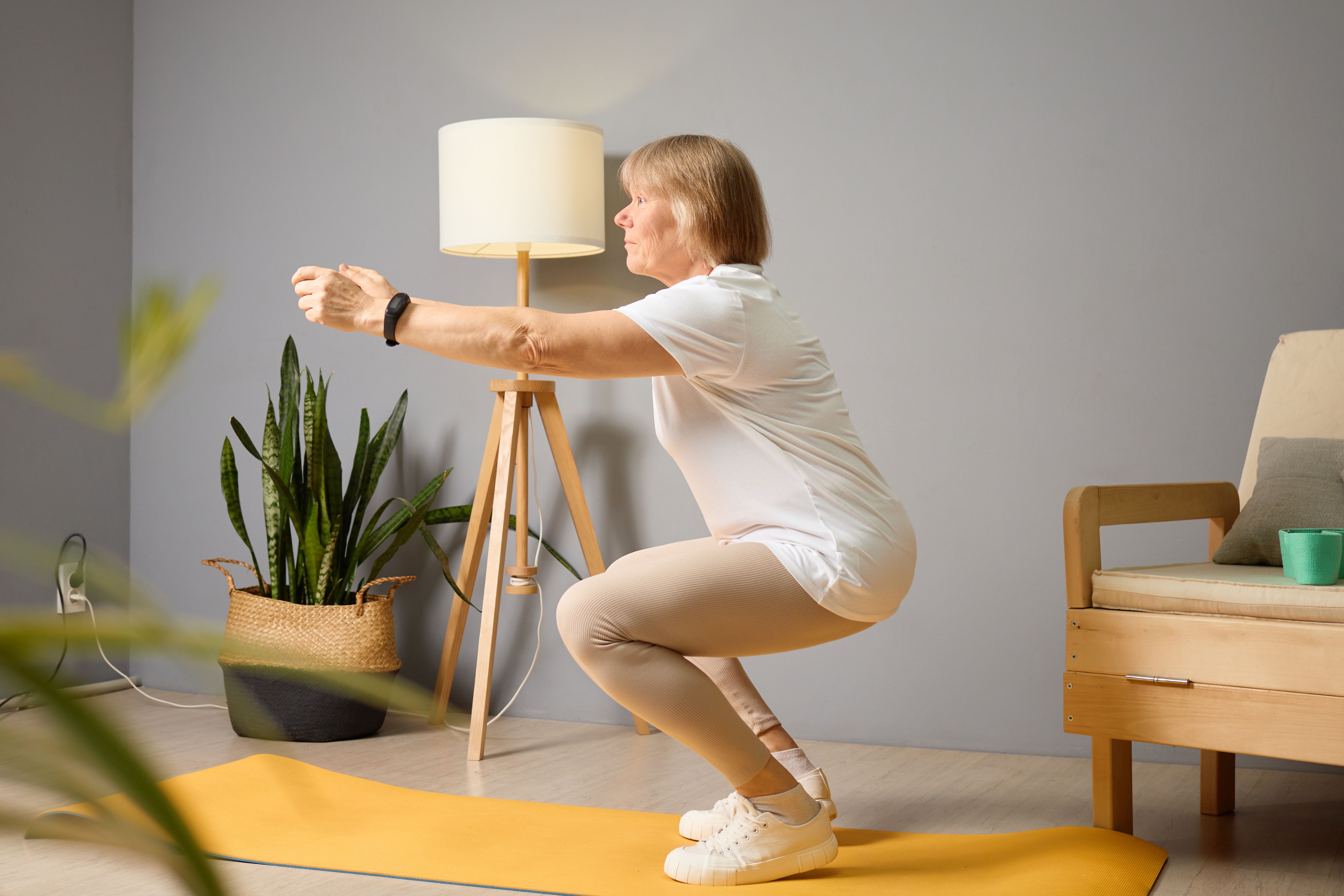 Senior woman performing squats indoors in a well-lit living room on a yoga mat, promoting fitness and health in a comfortable environment
