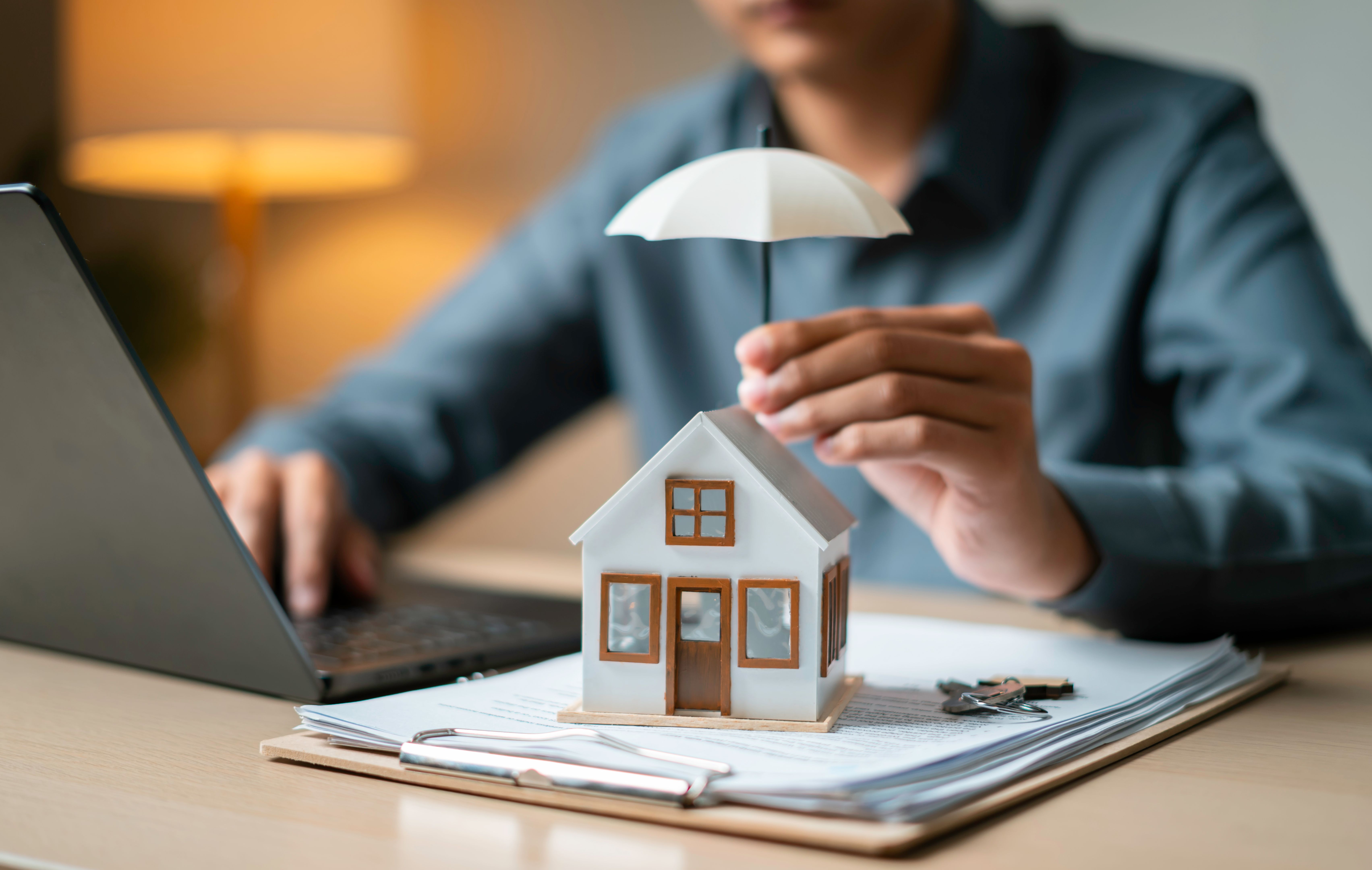 Close-up of a tiny house shielded by an umbrella atop loan documents next to keys and a laptop, representing property coverage. Close-up of a tiny house shielded by an umbrella atop loan documents next to keys and a laptop, representing property coverage.