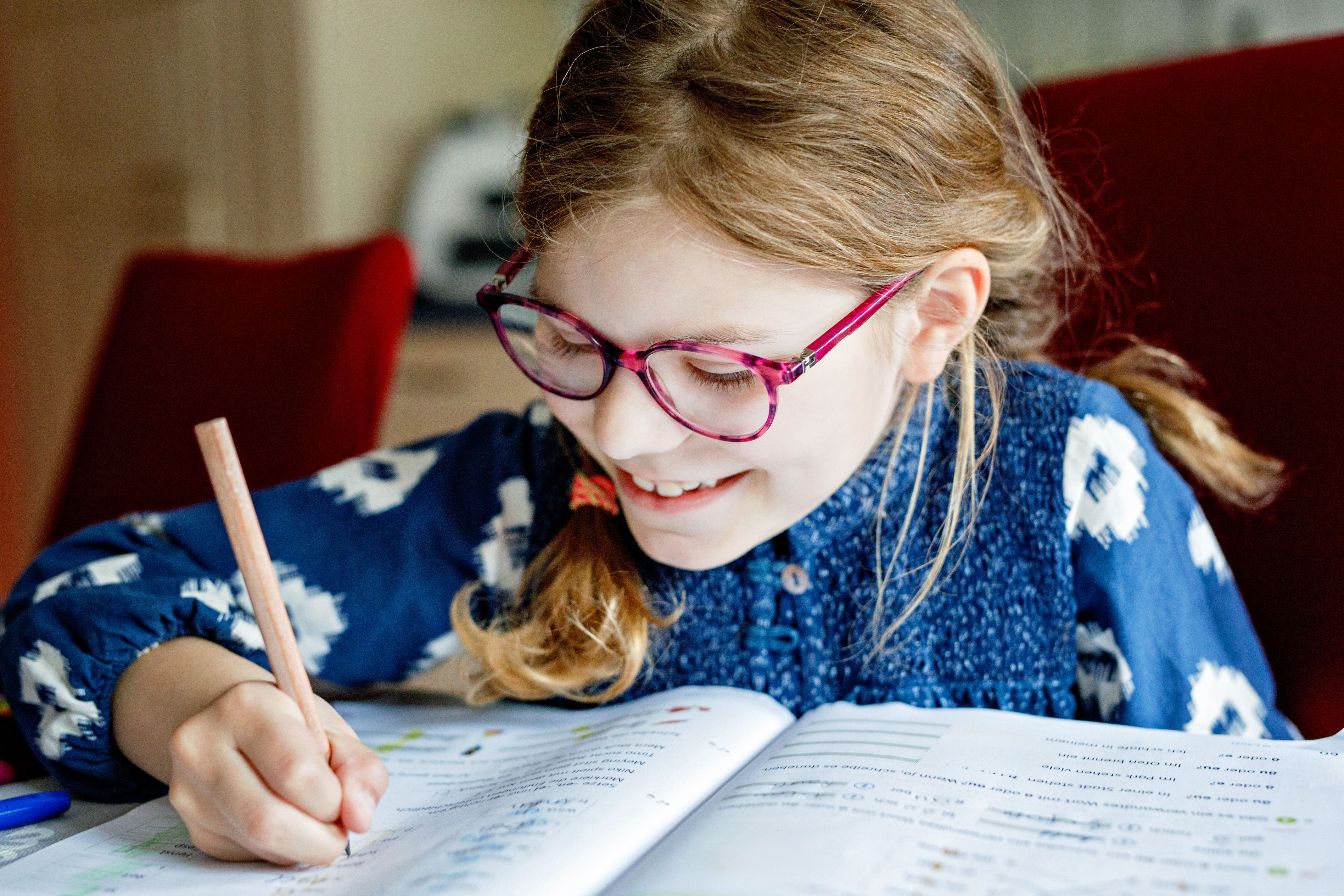 Cute elementary school girl sitting at a desk at home, writing in her workbook and doing school homework. Focused child learning and studying indoors. Concept of education, childhood, home learning.