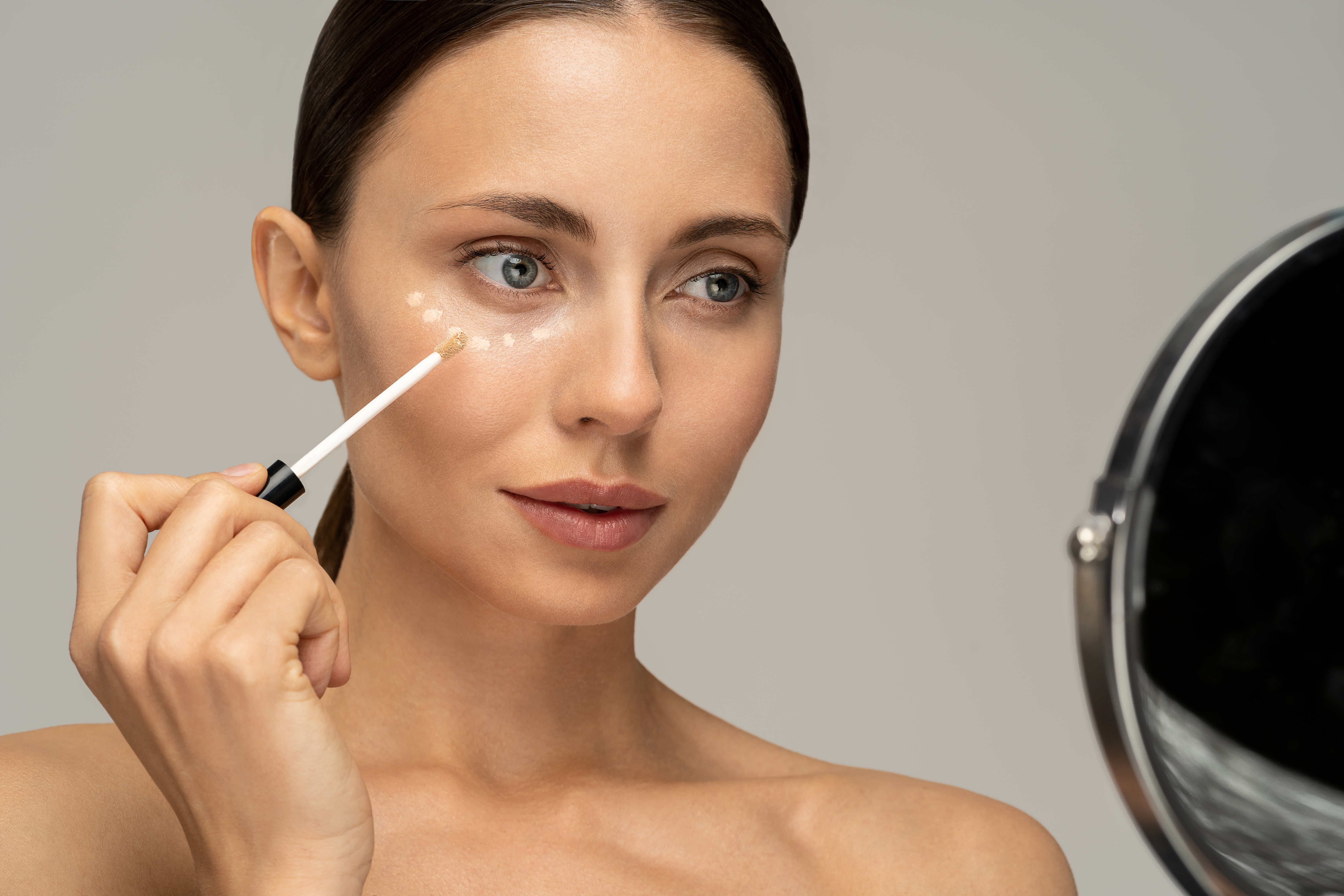 Woman with natural makeup applying corrector on flawless fresh skin, doing make up, looking at mirror. Girl after shower put concealer under eye area