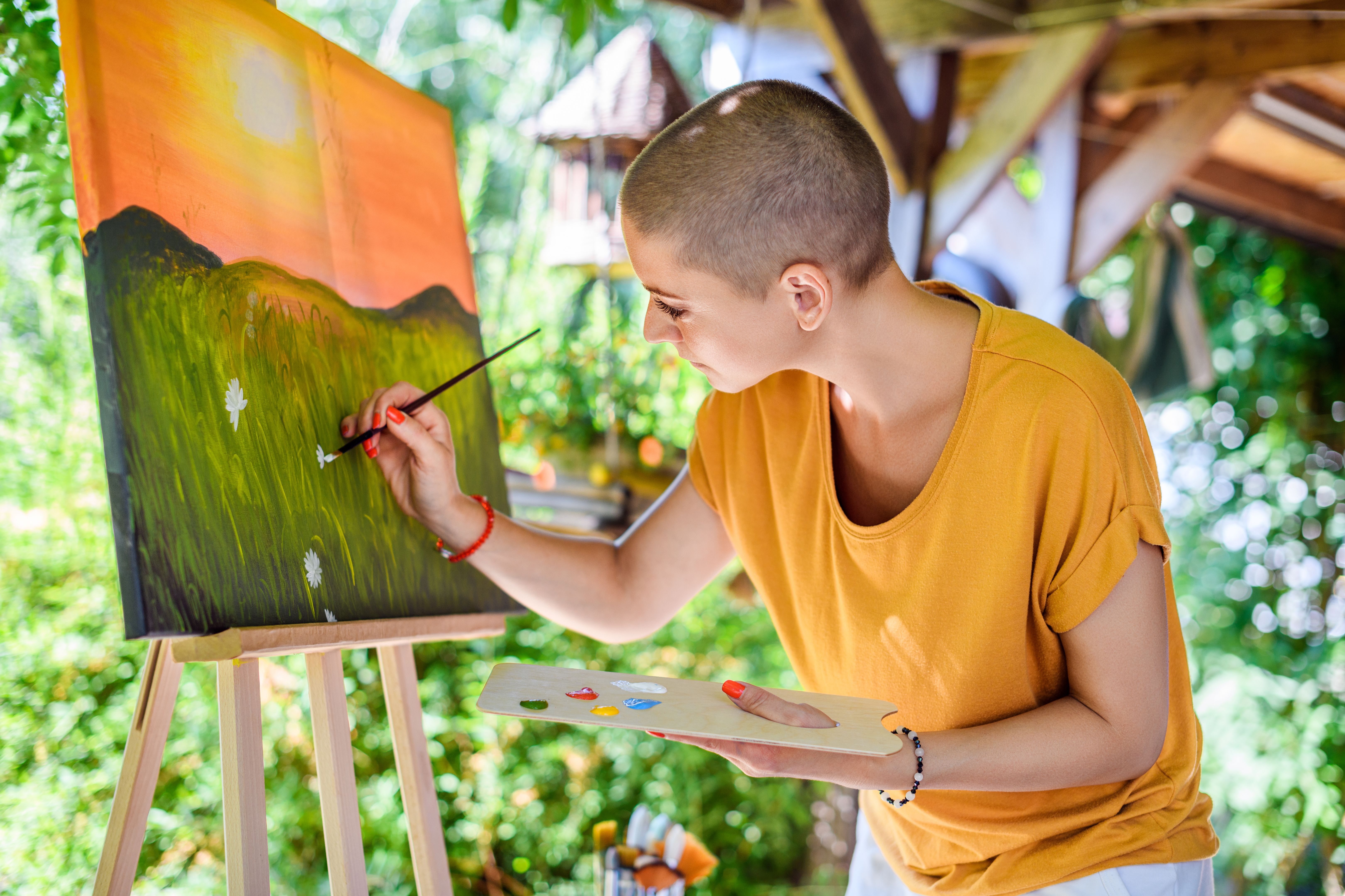Young female artist working on her art canvas painting outdoors in her garden. Cancer survivor, mindfulness, art therapy, creativity concept.