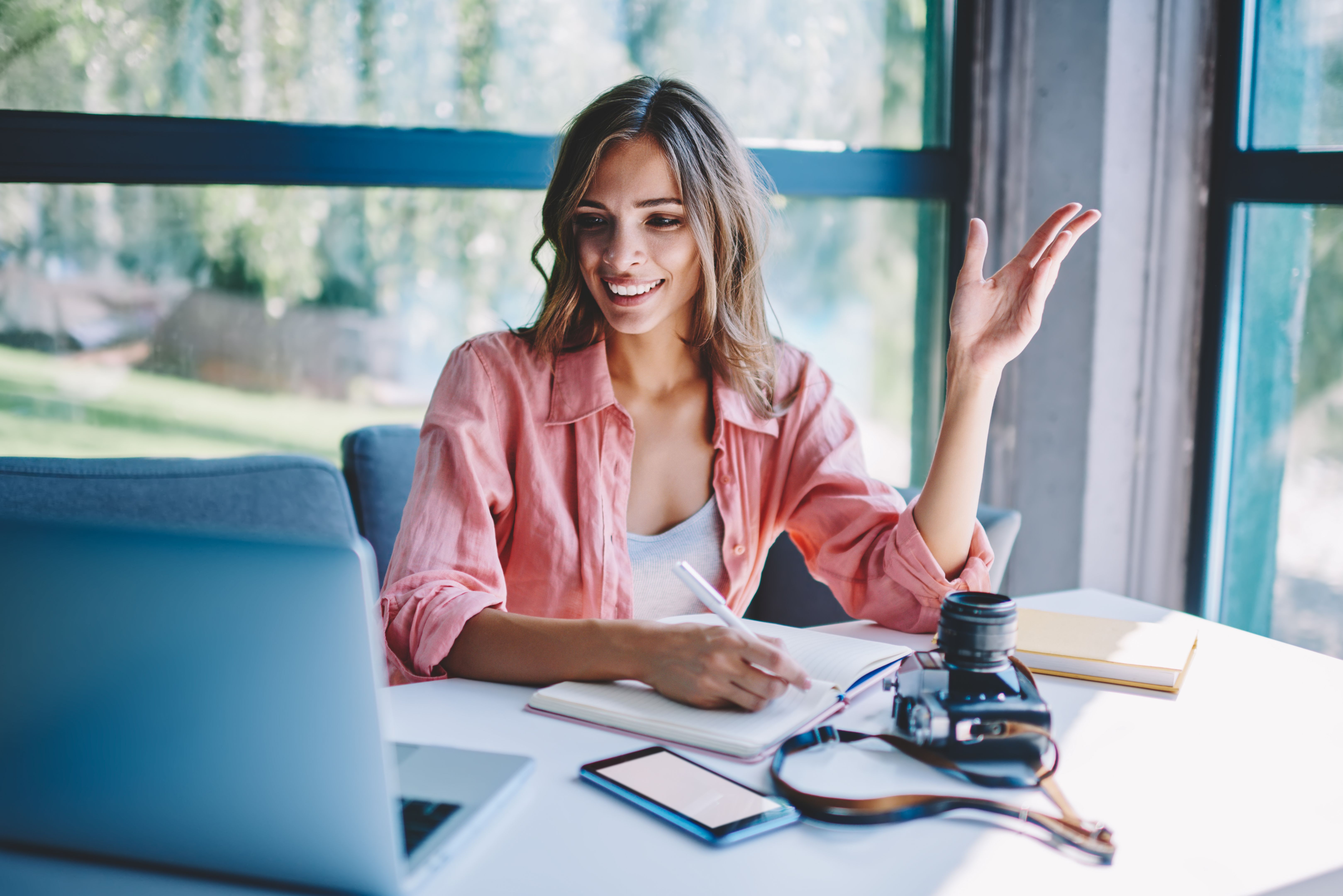 Happy young woman watching successful business webinar on website and making notes in notepad using laptop with wireless internet in cafe.Positive hipster student reading news and writing text