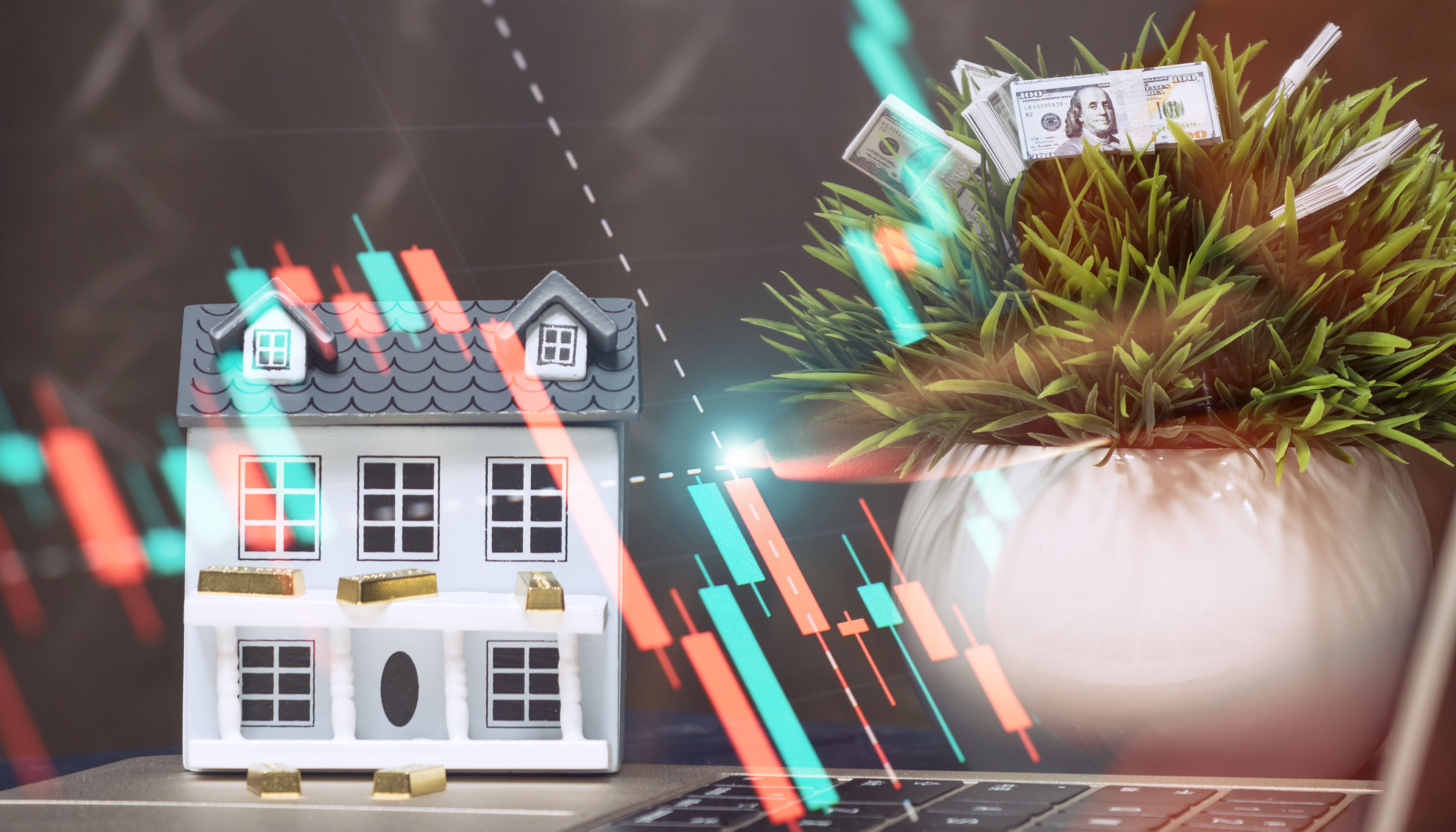 Market analysis and real estate investment workspace with miniature house, gold bars, green plant filled with money, candlestick chart on laptop
