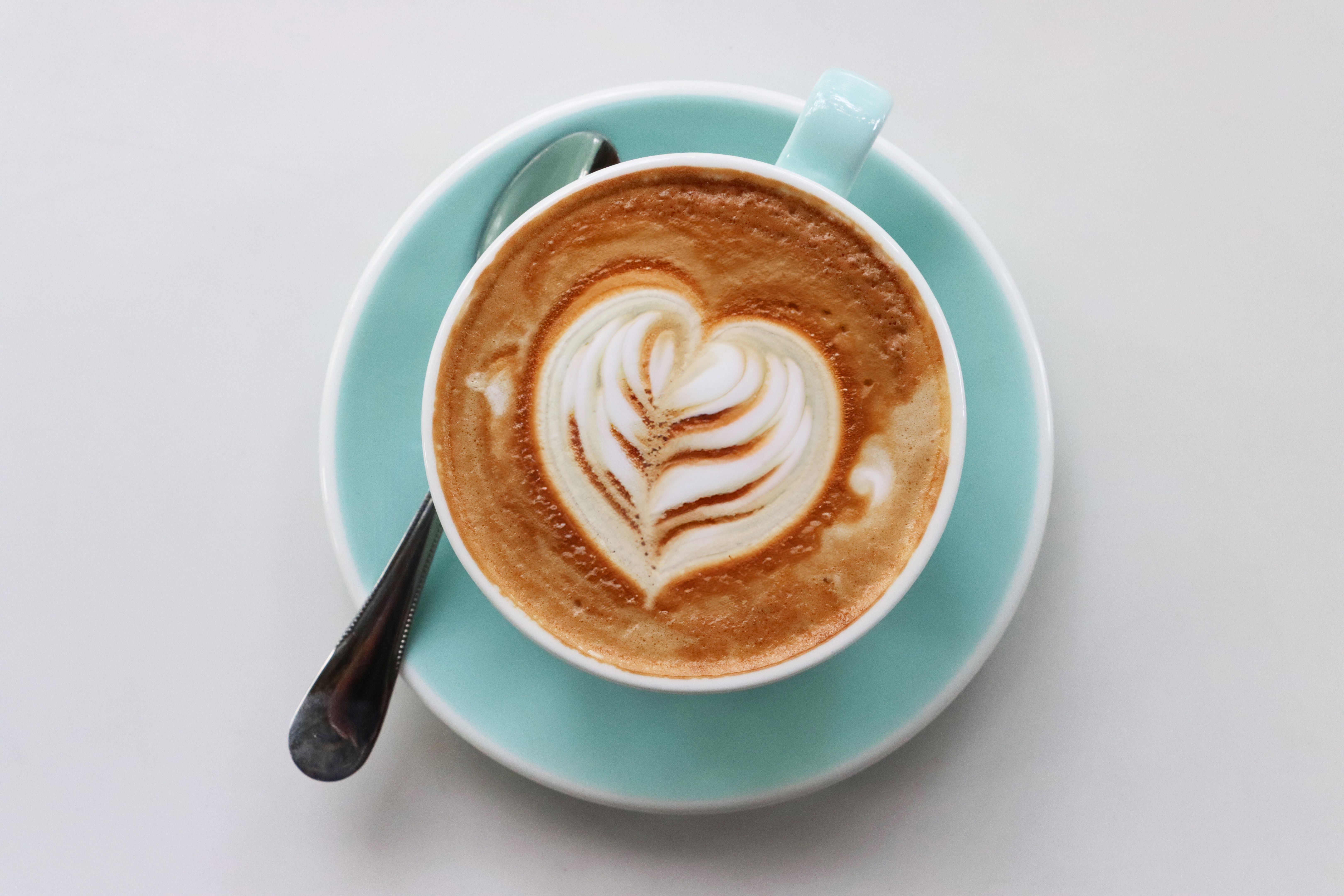 Close-up image of white foam coffee latte art, light teal blue cup and saucer with metal teaspoon, heart shape cappuccino froth art design, coffee shop cafe table, frothy latte hot drink, white background, elevated view, copy space Close-up image of white foam coffee latte art, light teal blue cup and saucer with metal teaspoon, heart shape cappuccino froth art design, coffee shop cafe table, frothy latte hot drink, white background, elevated view, copy space
