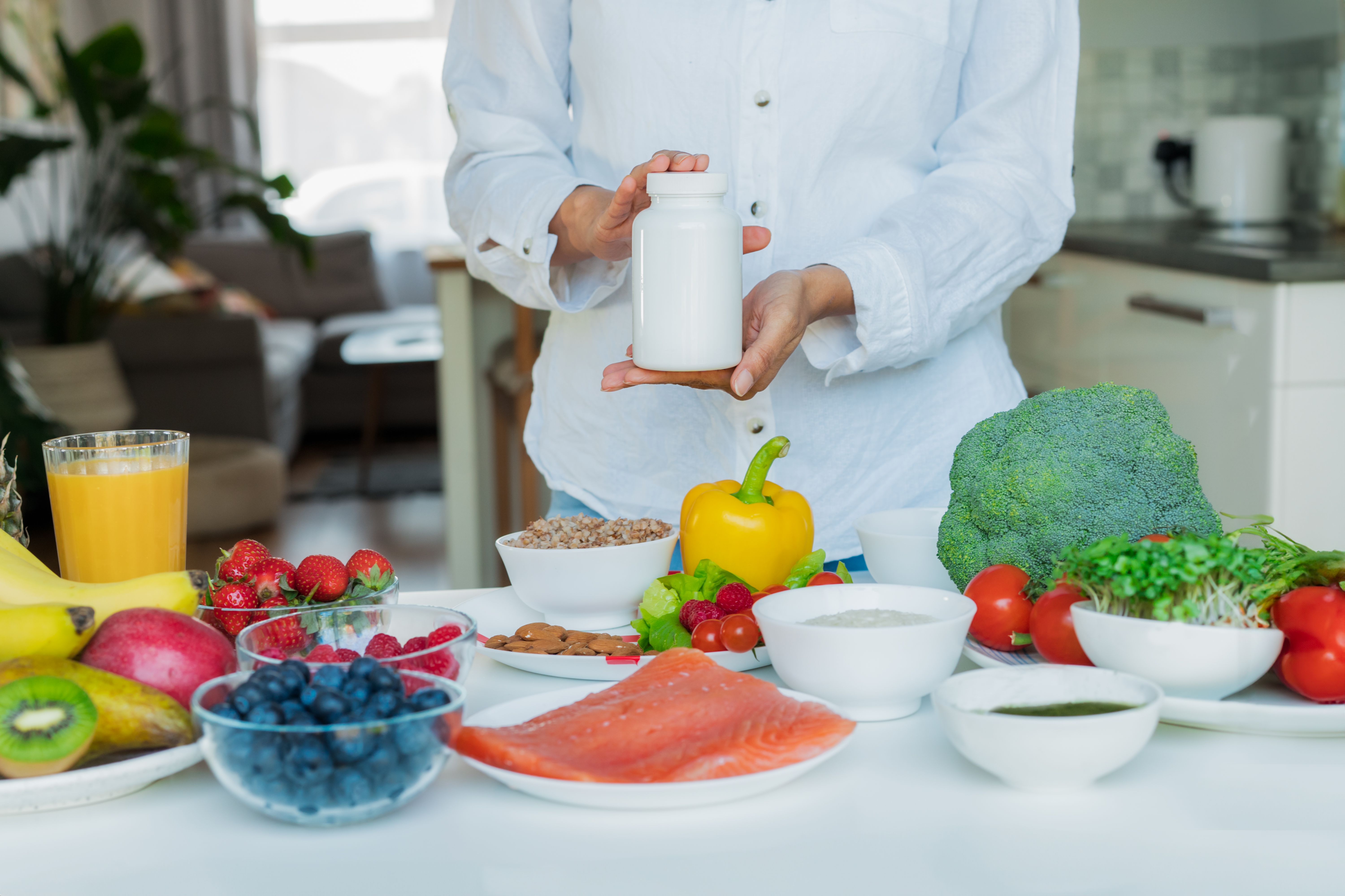 Focus on female hand holding white mockup jar with nutritional supplements on table with fresh food ingredients. Concept of Balanced Diet. Supplementing diet with missing vitamins. Dietary supplement