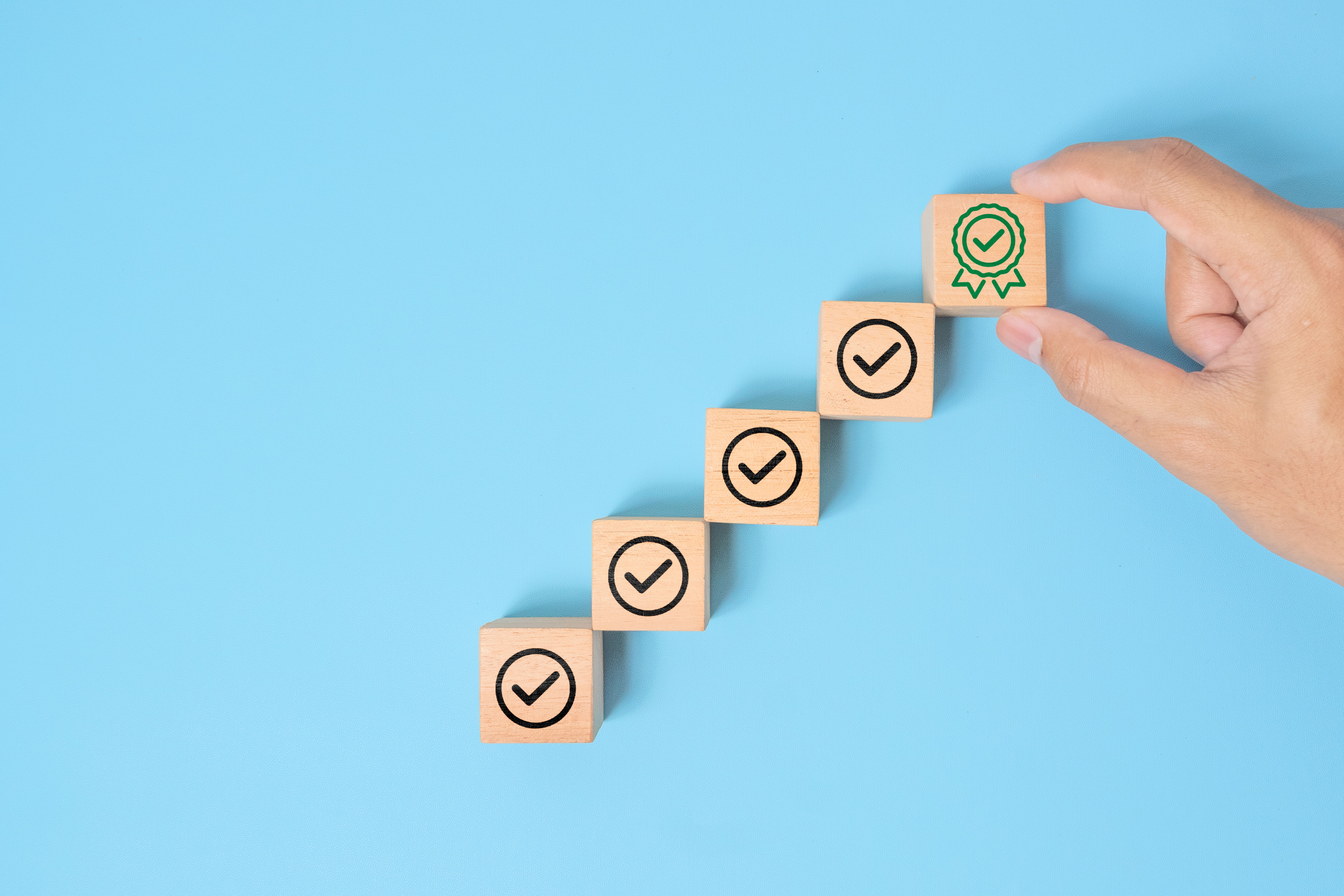 A hand is placing the final wooden block with a green certificate checkmark, symbolizing achievement, quality assurance and completion, against a plain pastel blue background