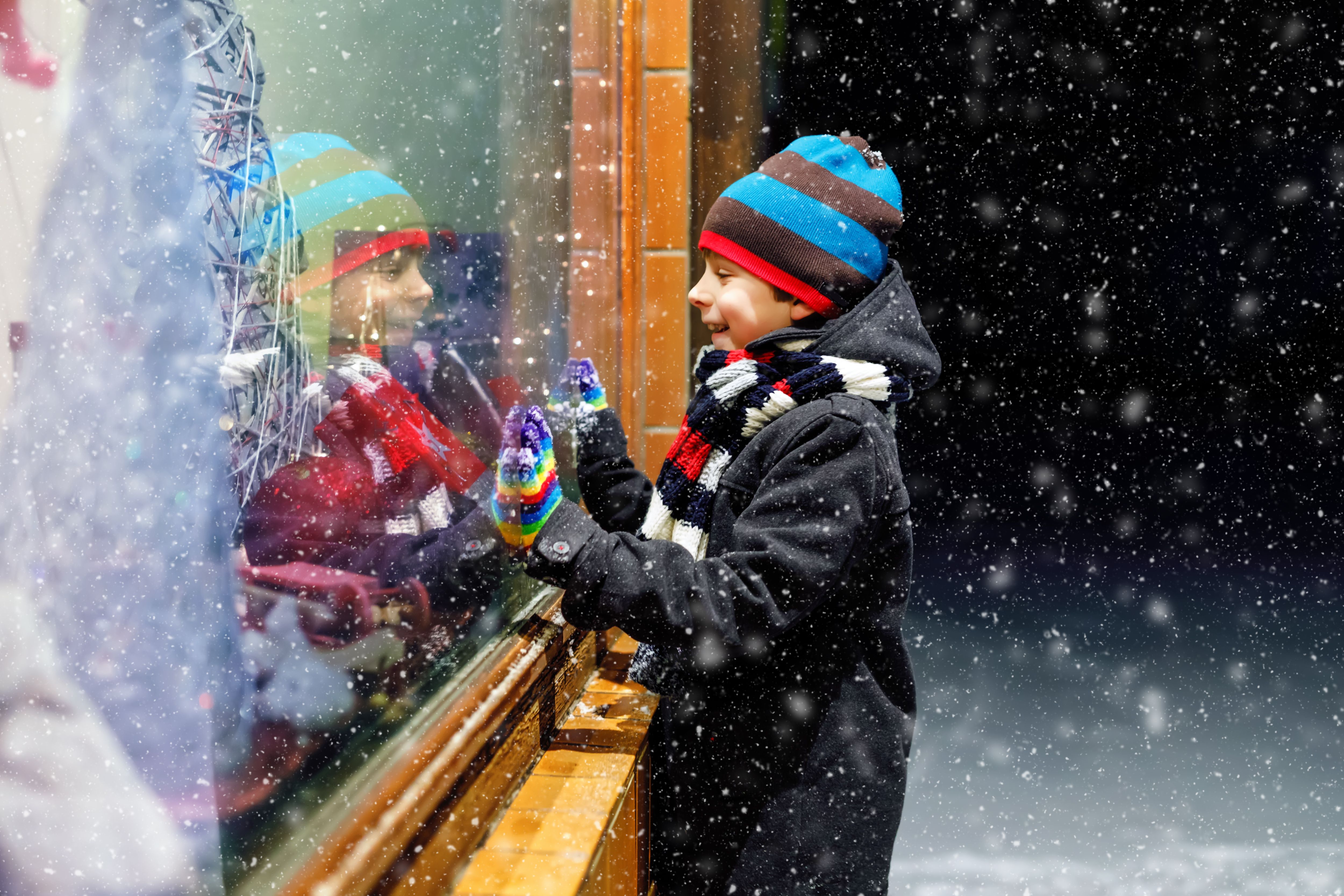 Cute little healthy school kid boy on Christmas market. Funny happy child in fashion winter clothes making window shopping decorated with gifts, xmas tree. Snow falling down, snowfall