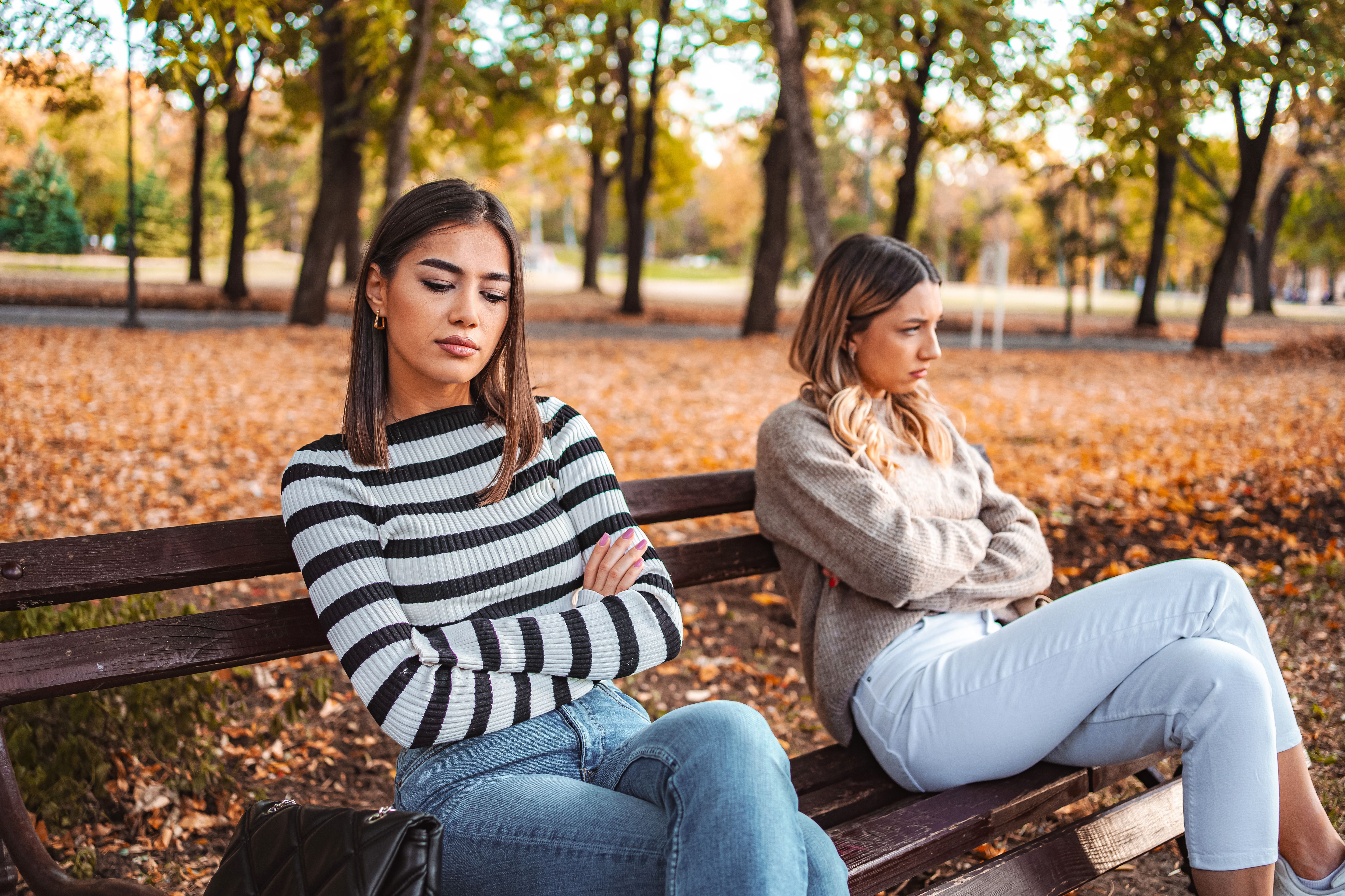 Two young women having an argument while sitting on a bench in a park during autumn Two young women having an argument while sitting on a bench in a park during autumn