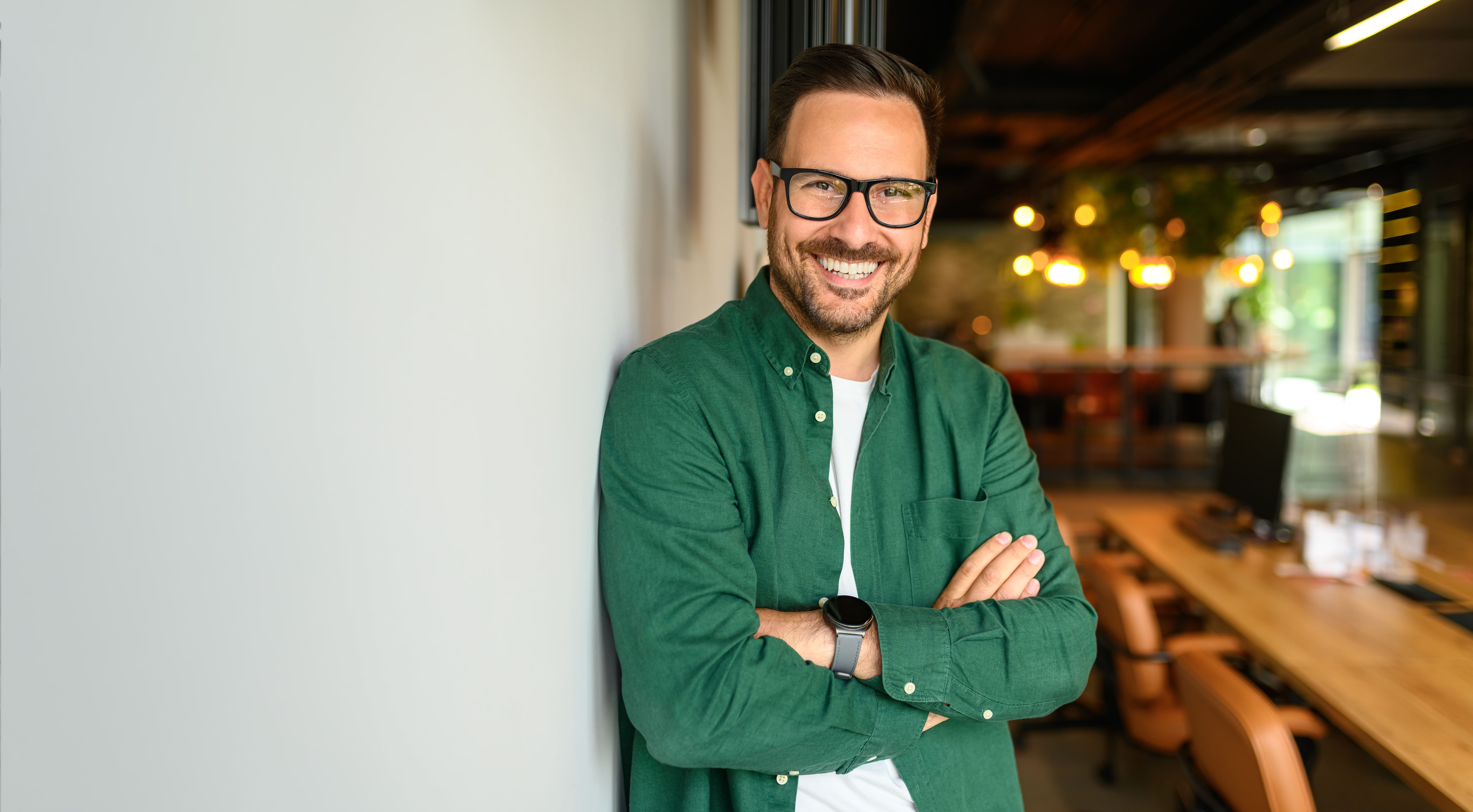 Portrait of confident businessman in glasses and with arms crossed leaning on white wall in office
