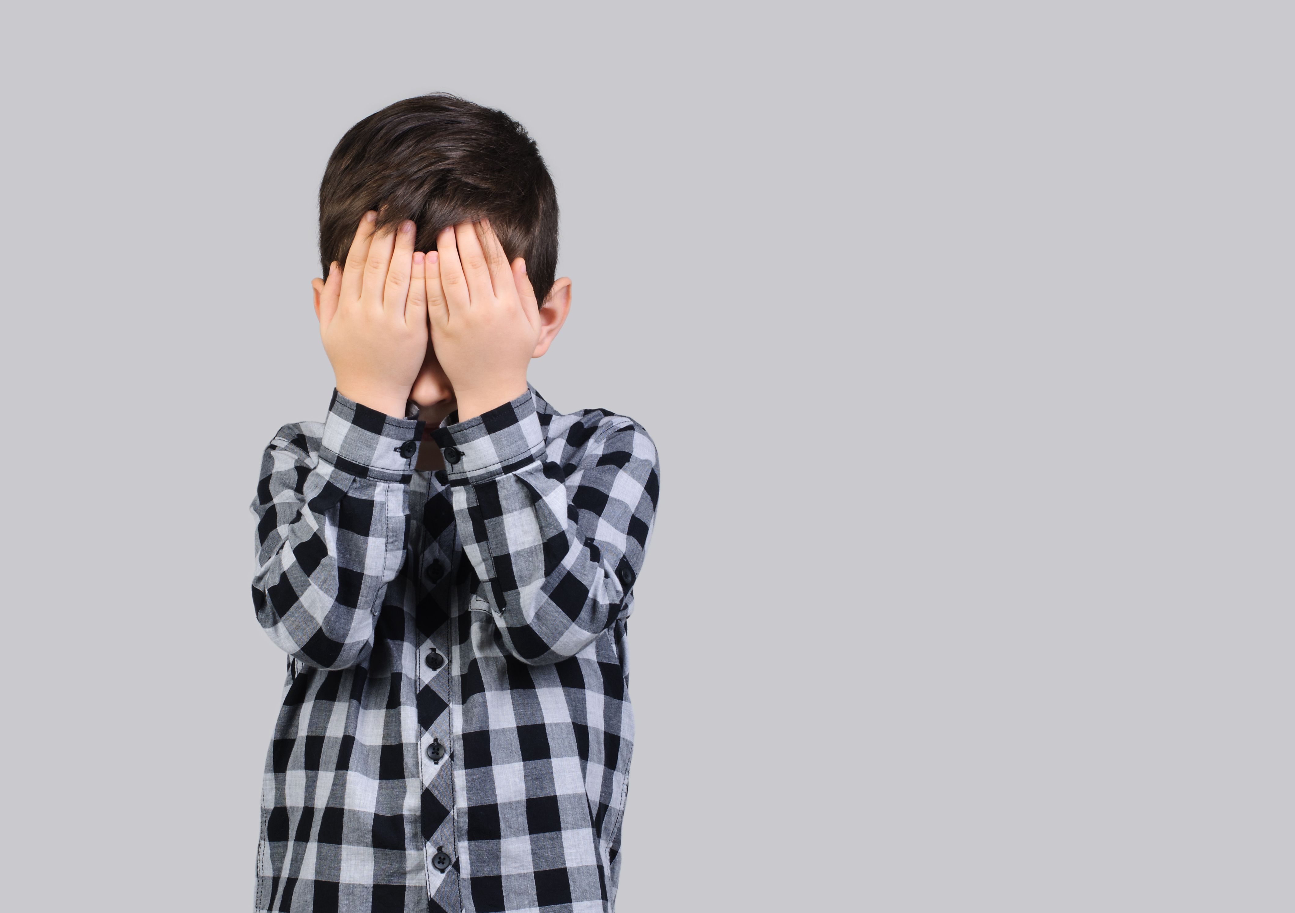 Unhappy schoolboy, 6-year-old boy covered his face with his hands on studio gray isolated background.
