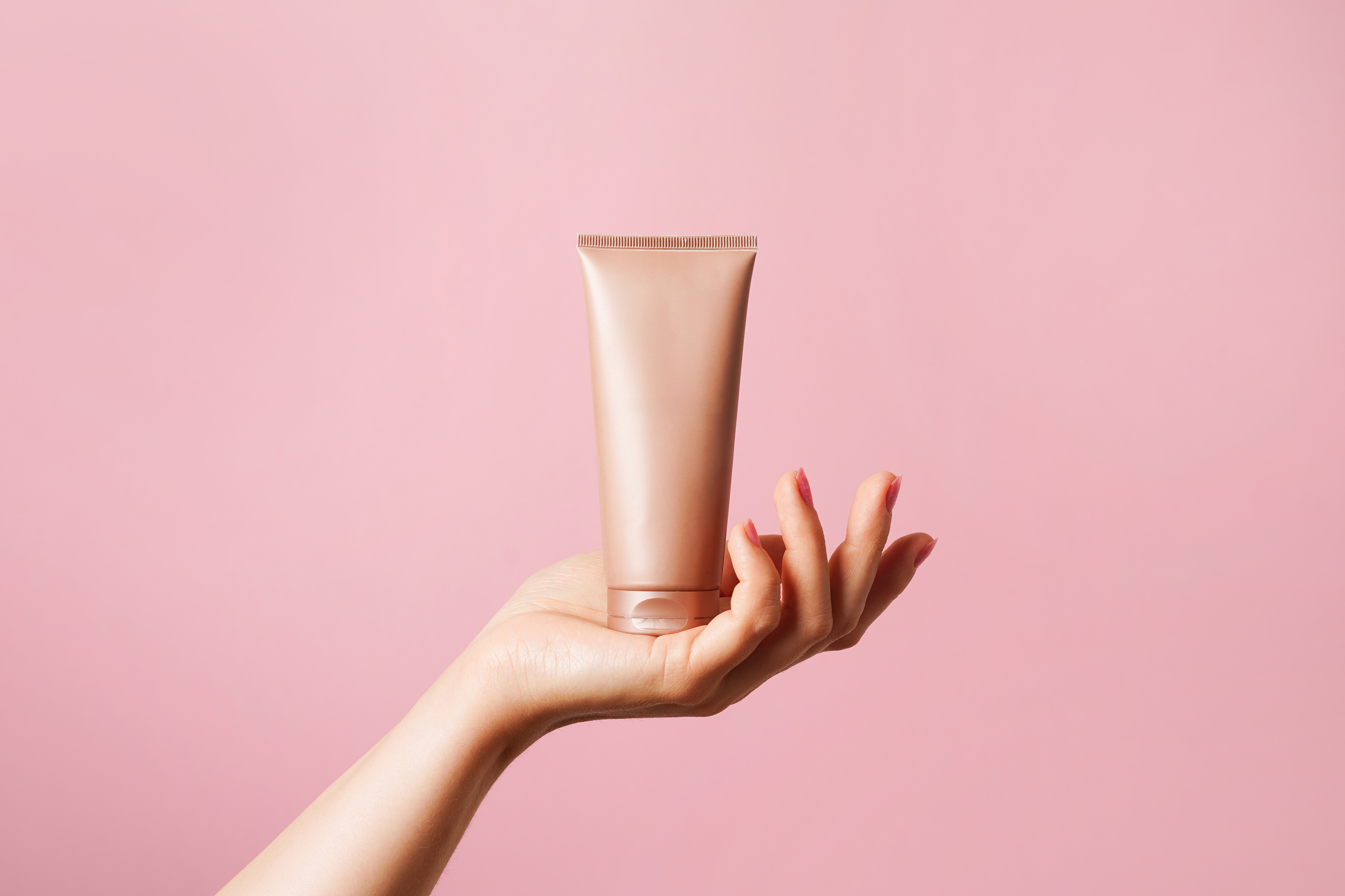 Woman hand holding unbranded beige tube with cosmetic cream on pink background. skin care concept, copy space