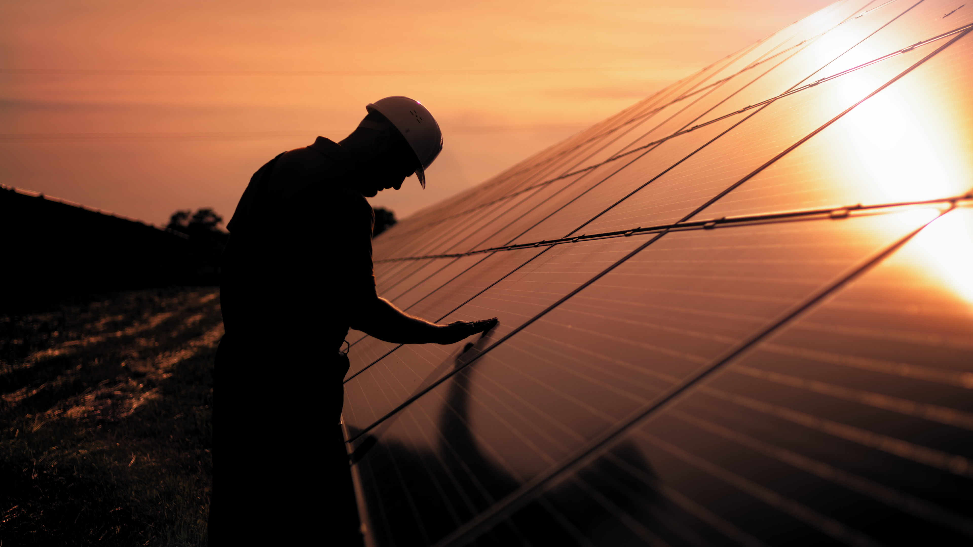 Assistance technical worker in uniform is checking an operation and efficiency performance of photovoltaic solar panels. Unidentified solar power engineer touches solar panels with his hand at sunset