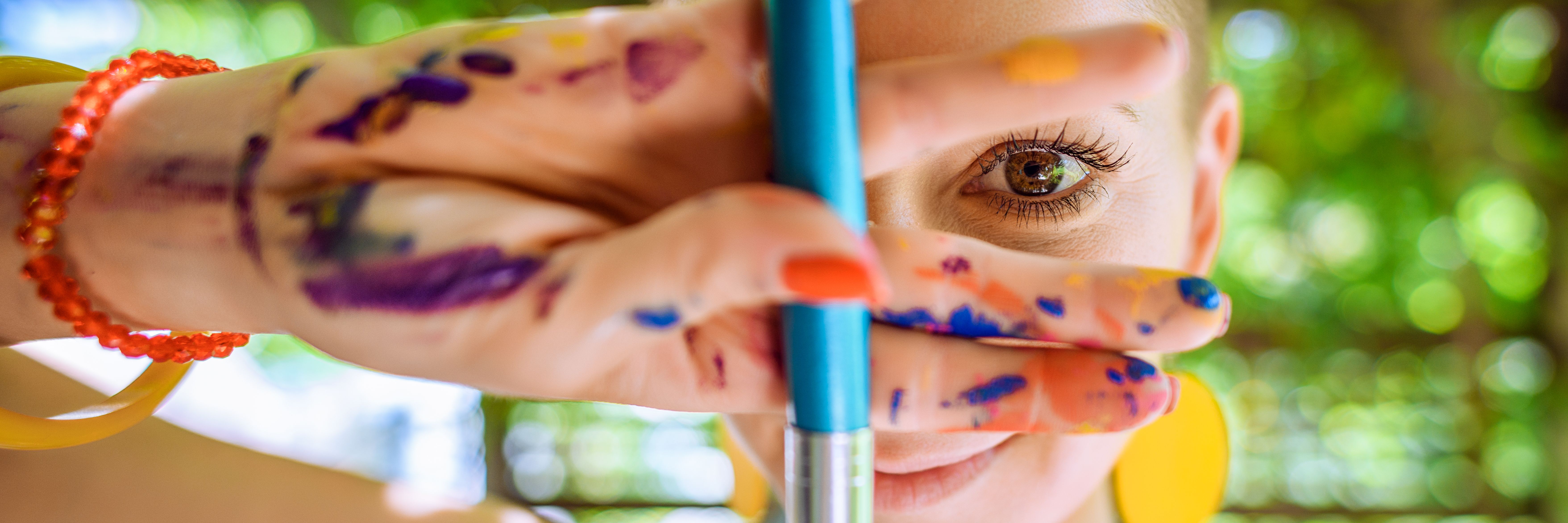 Playful portrait of a young gorgeous female painter artist, with hands covered in paint, looking and smiling at camera through her fingers. Creativity and individuality banner.