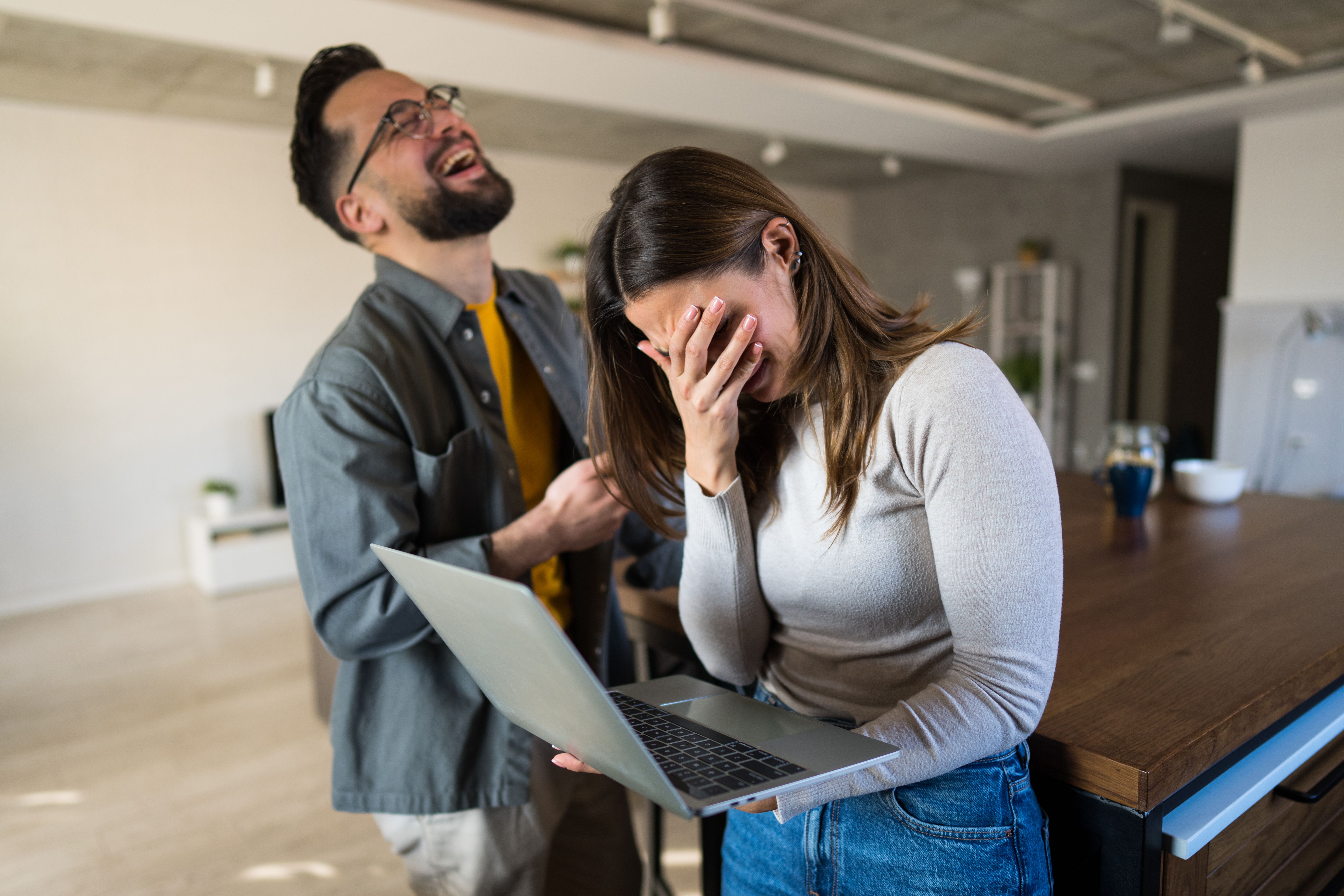 Young woman feeling ashamed while reading something on laptop with her husband laughing Young woman feeling ashamed while reading something on laptop with her husband laughing