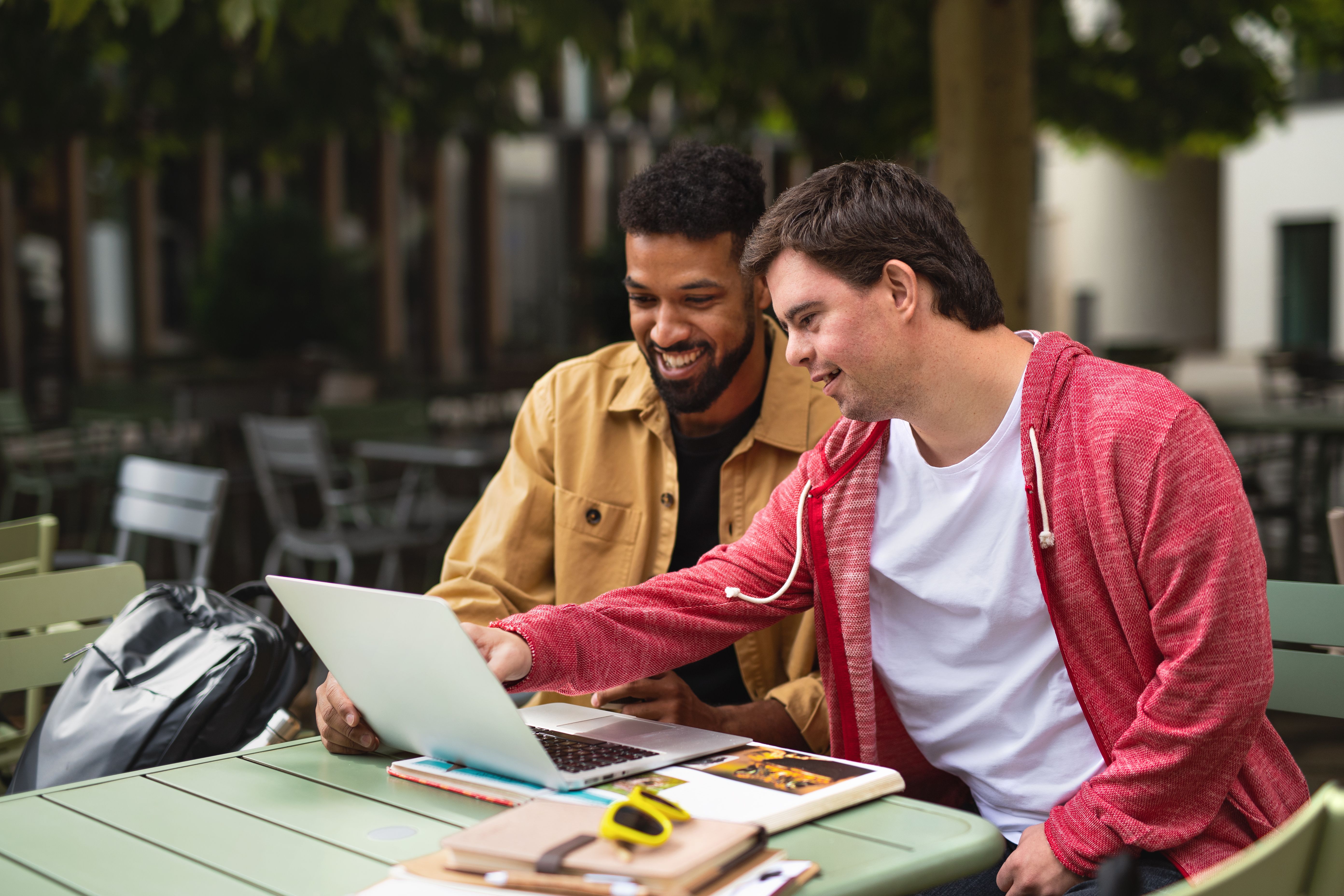 Young man with Down syndrome with his mentoring friend sitting outdoors in cafe using laptop.