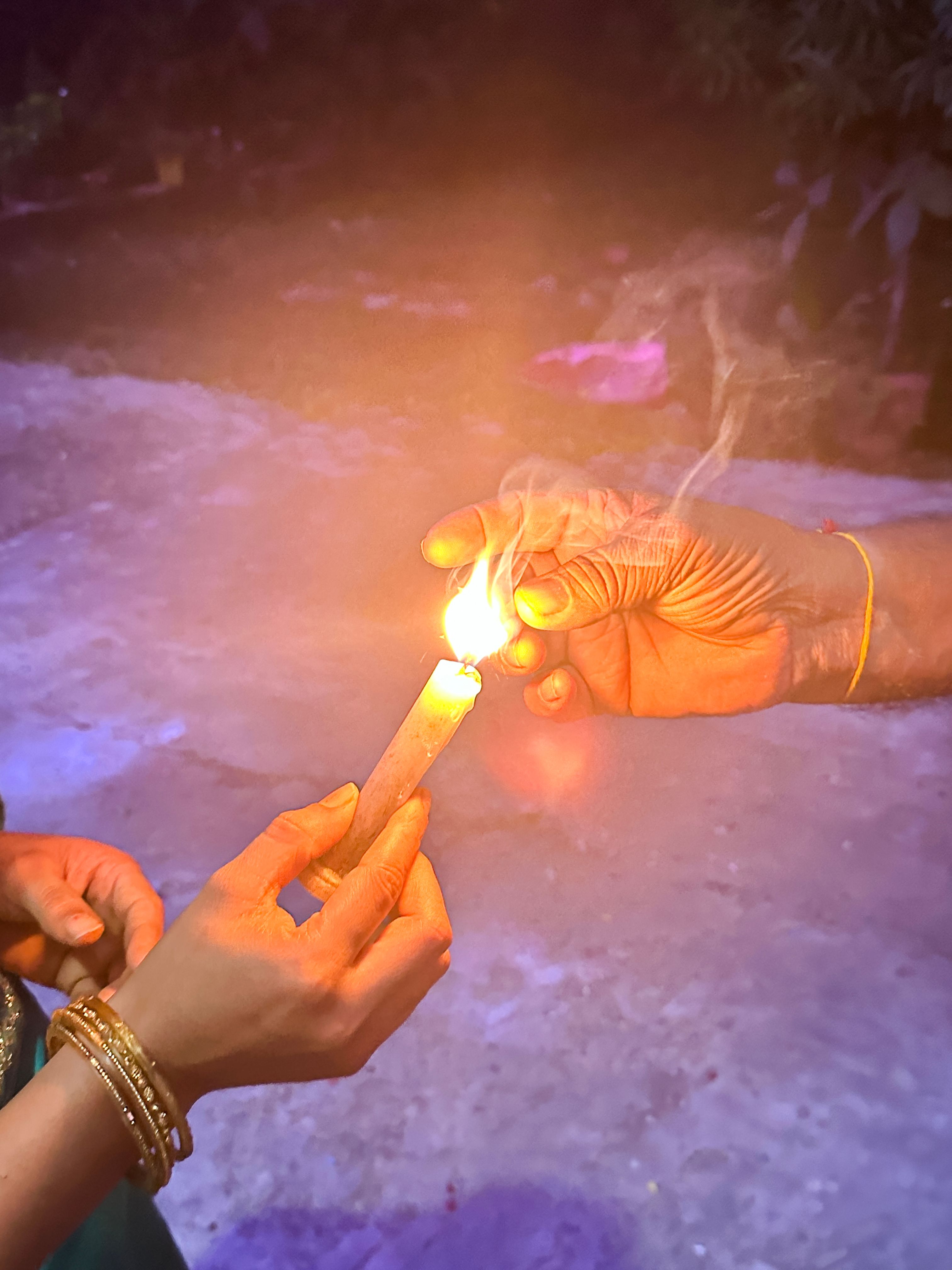 Close-up image of unrecognisable woman wearing gold bangles, holding candle used to light mitti clay diya, seconf unrecognisable person extinguishing flame between fingers and thumb, Diwali festival of lights celebration, focus on foreground