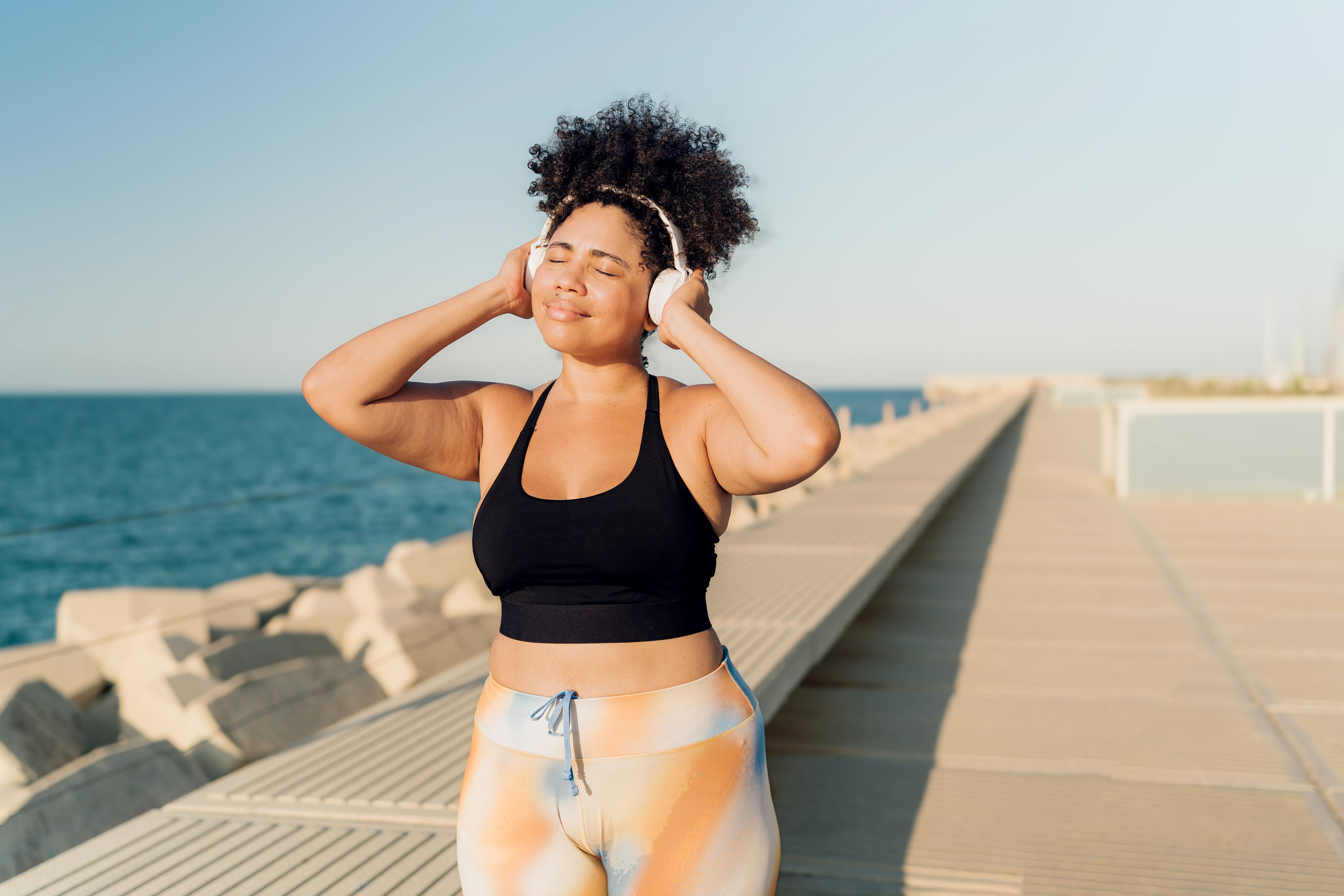Plus-size Hispanic sportswoman enjoying music with headphones on pier by the sea