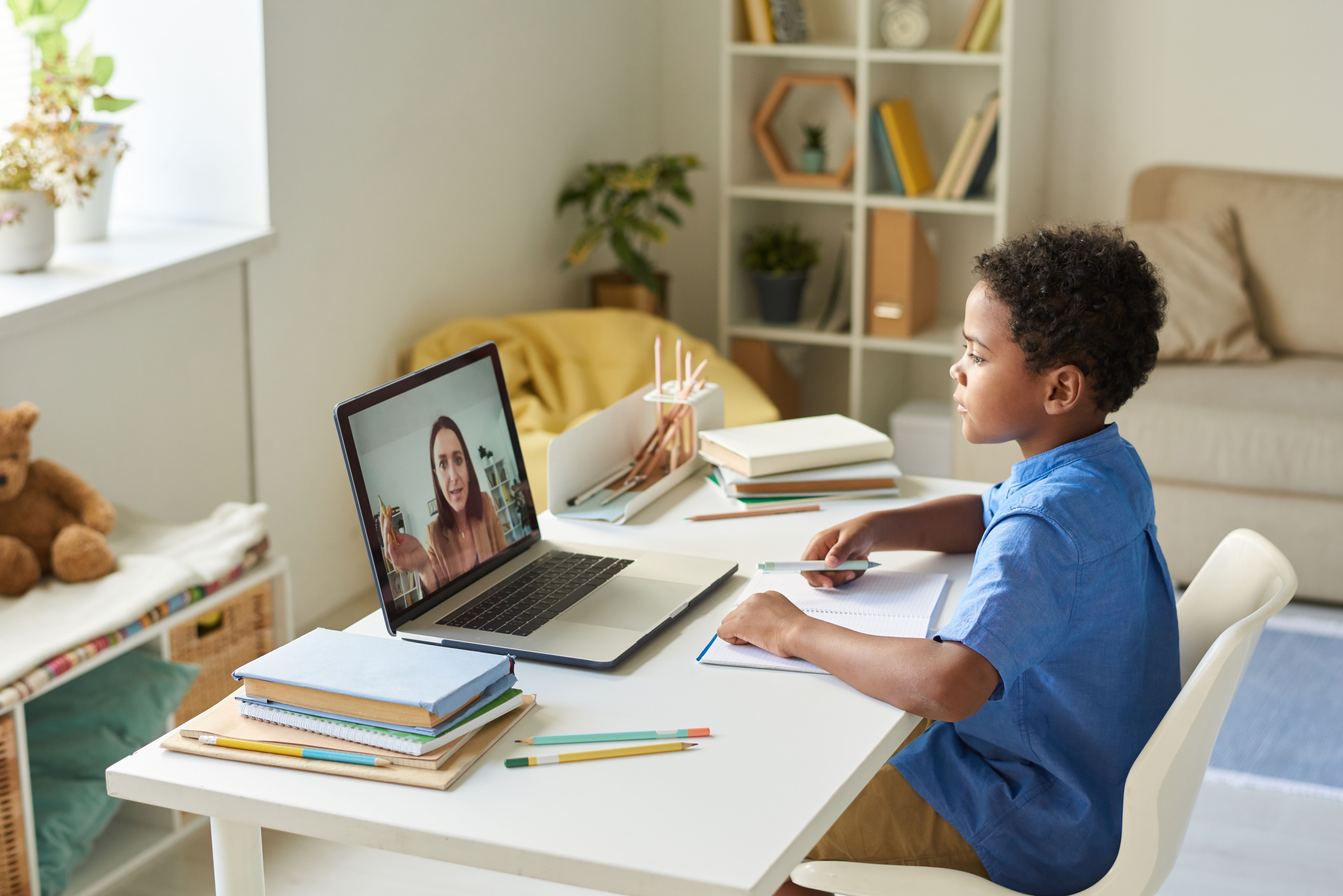 Focused black schoolboy with curly hair sitting at desk and using laptop while listening to tutor during video conference Focused black schoolboy with curly hair sitting at desk and using laptop while listening to tutor during video conference