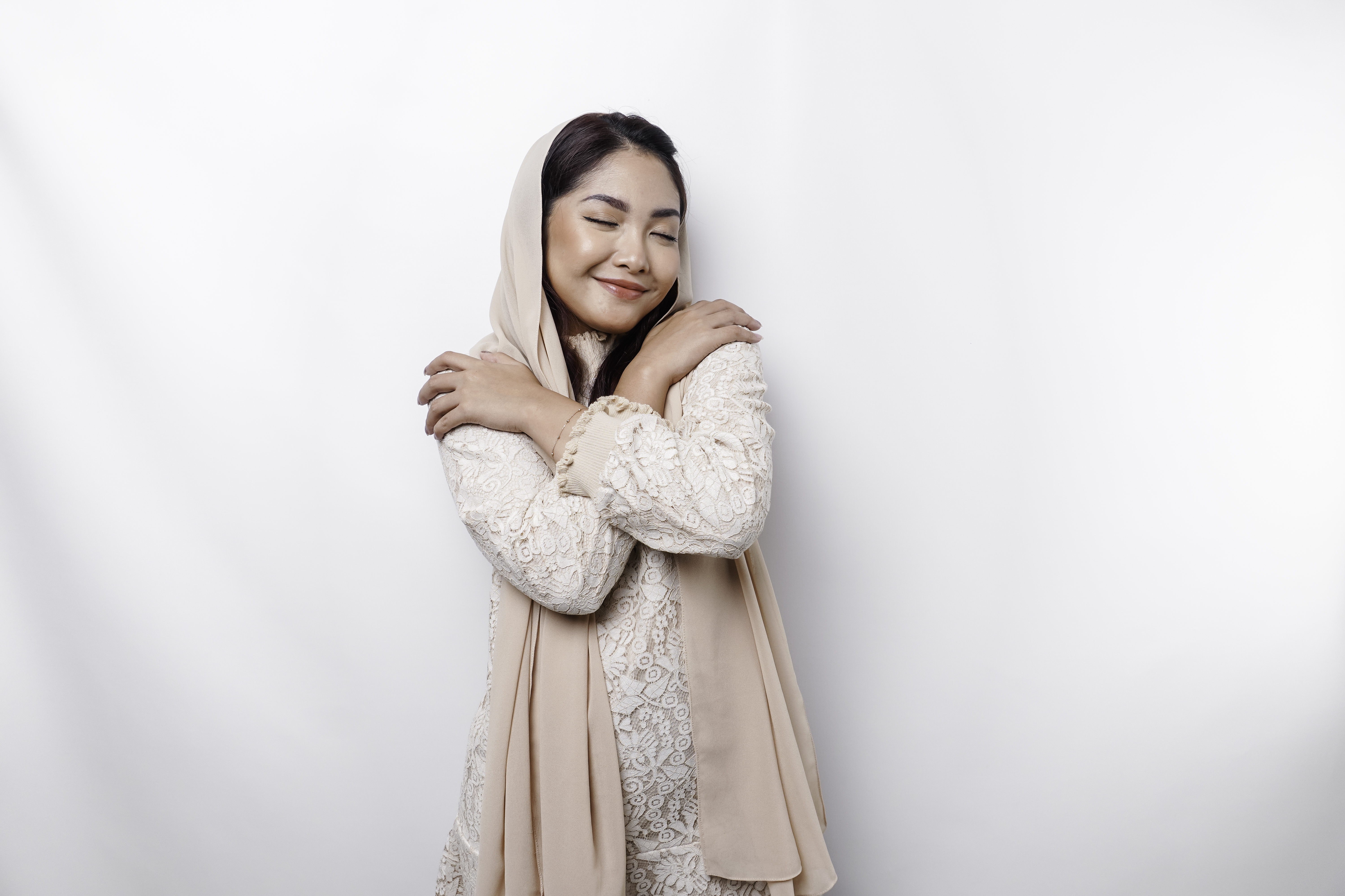 Young beautiful Asian Muslim woman wearing a headscarf over white background hugging herself happy and positive, smiling confident. Self-love and self-care