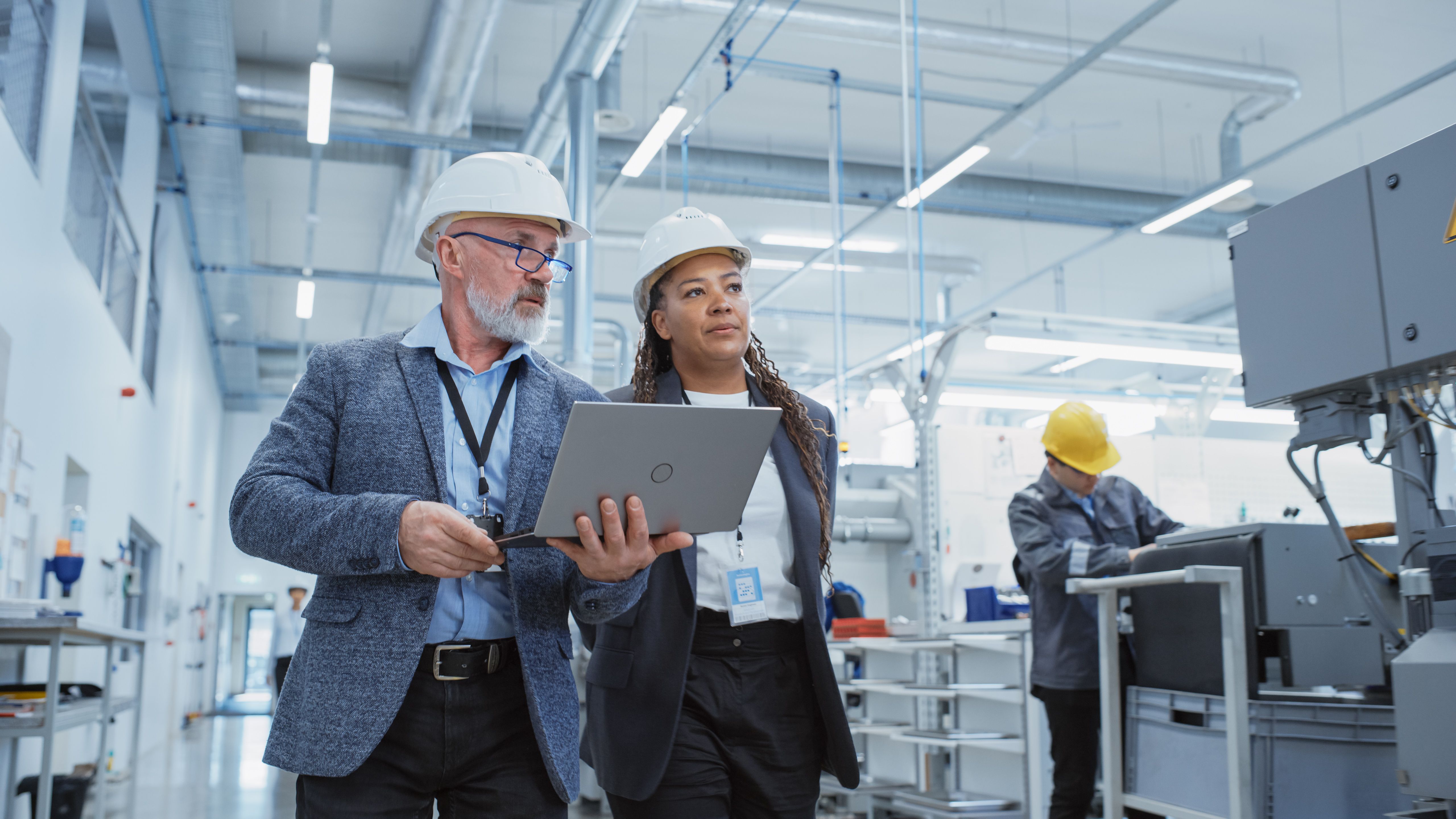 Two Professional Heavy Industry Engineers Wearing Hard Hats at Factory. Walking and Discussing Industrial Machine Facility, Working on Laptop. African American Manager and Technician at Work. Two Professional Heavy Industry Engineers Wearing Hard Hats at Factory. Walking and Discussing Industrial Machine Facility, Working on Laptop. African American Manager and Technician at Work.