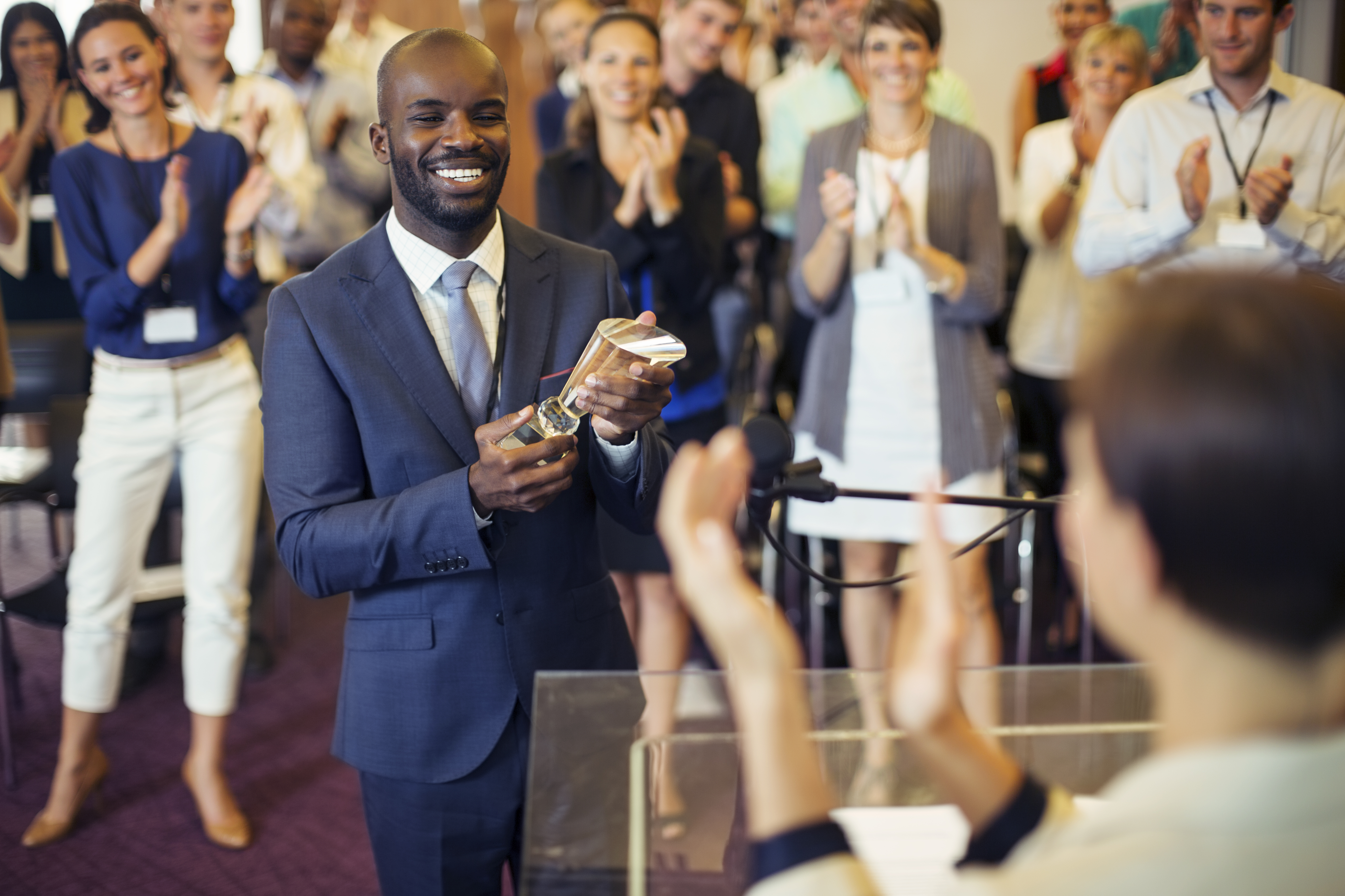Portrait of young man holding trophy, standing in conference room, smiling to applauding audience Portrait of young man holding trophy, standing in conference room, smiling to applauding audience
