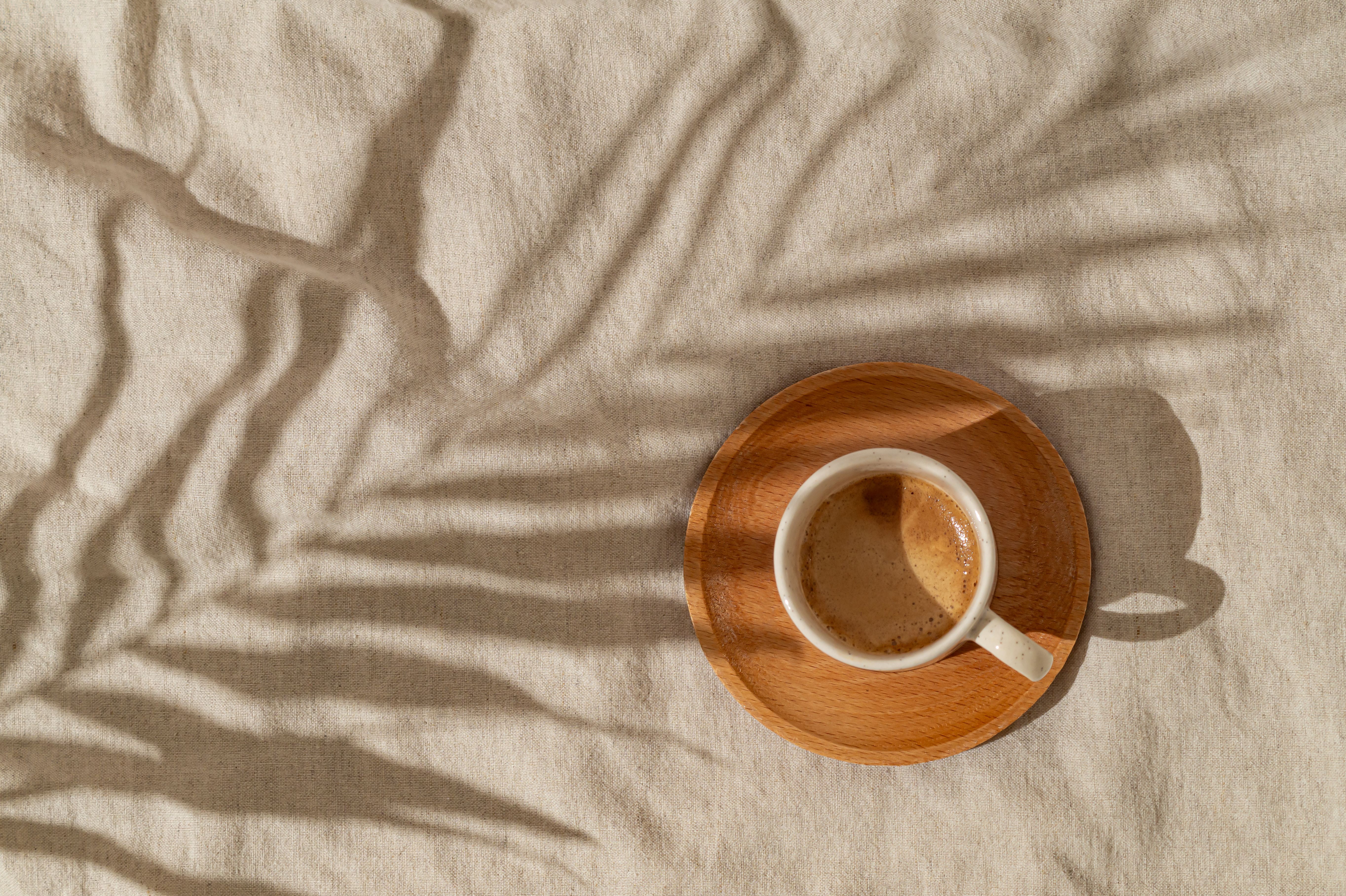 Summer day scene made with cup of coffee and tropical palm tree leaf shadow on sand color linen background. Sun and shadows. Minimal summer coffee concept. Flat lay style. Top view.