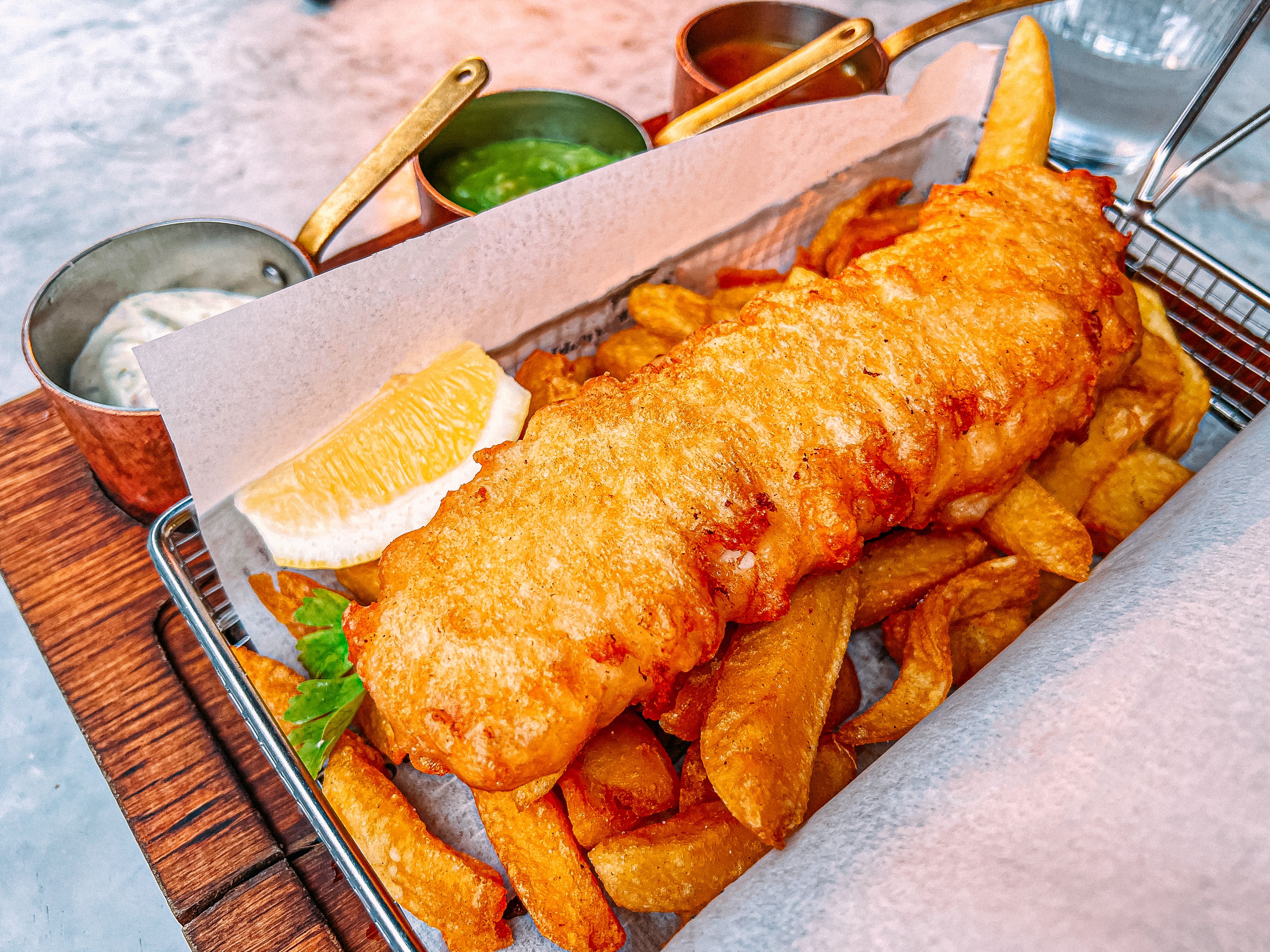 Close-Up Shot of a Plate of Delicious Perfectly Fried Fish and Chips Served at An Outdoor Restaurant with Tartar Sauce, Mushy Peas, and Curry Sauce