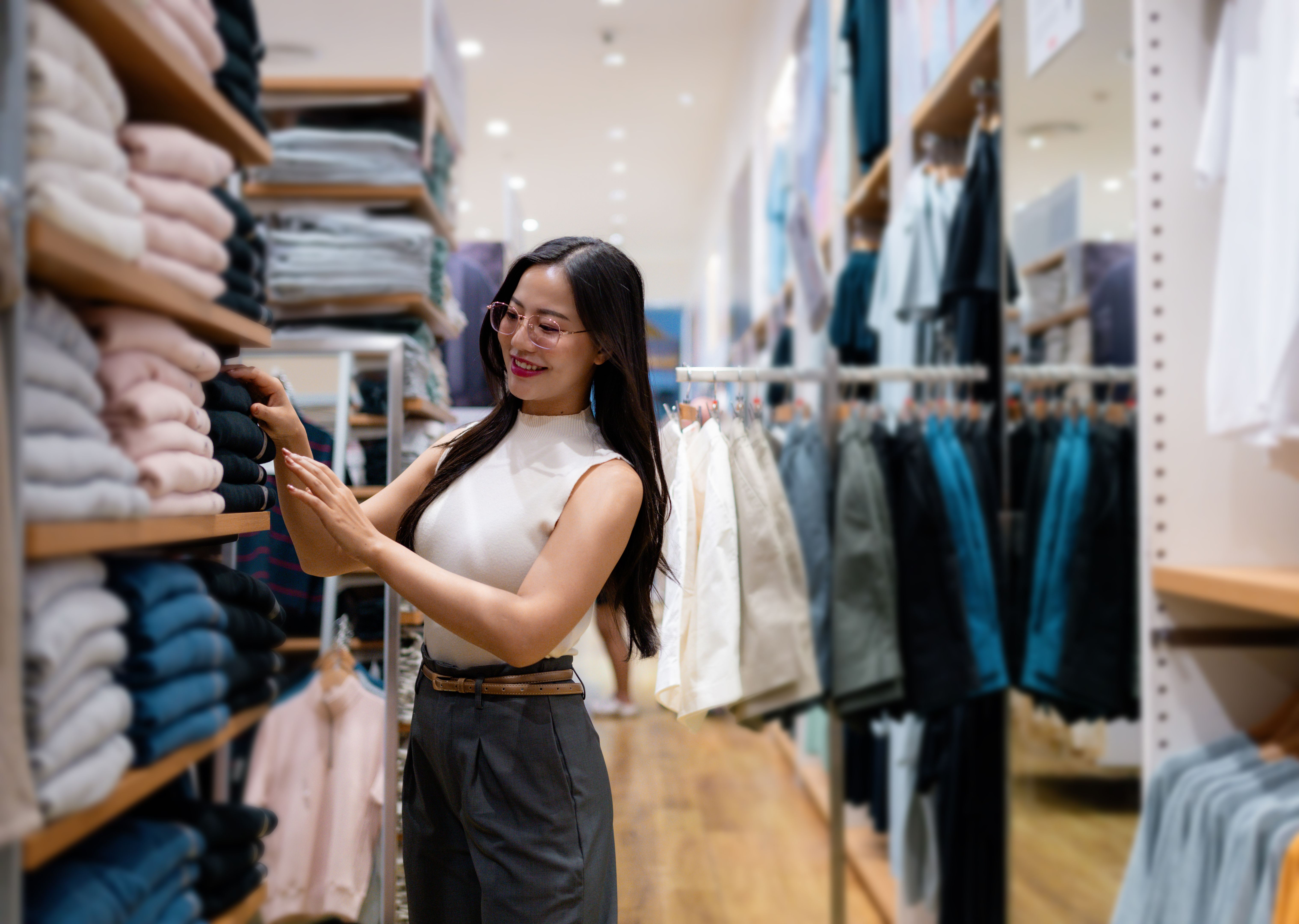 Smiling young woman selecting clothing from a shelf in a vibrant retail store, enjoying the process of finding stylish outfits