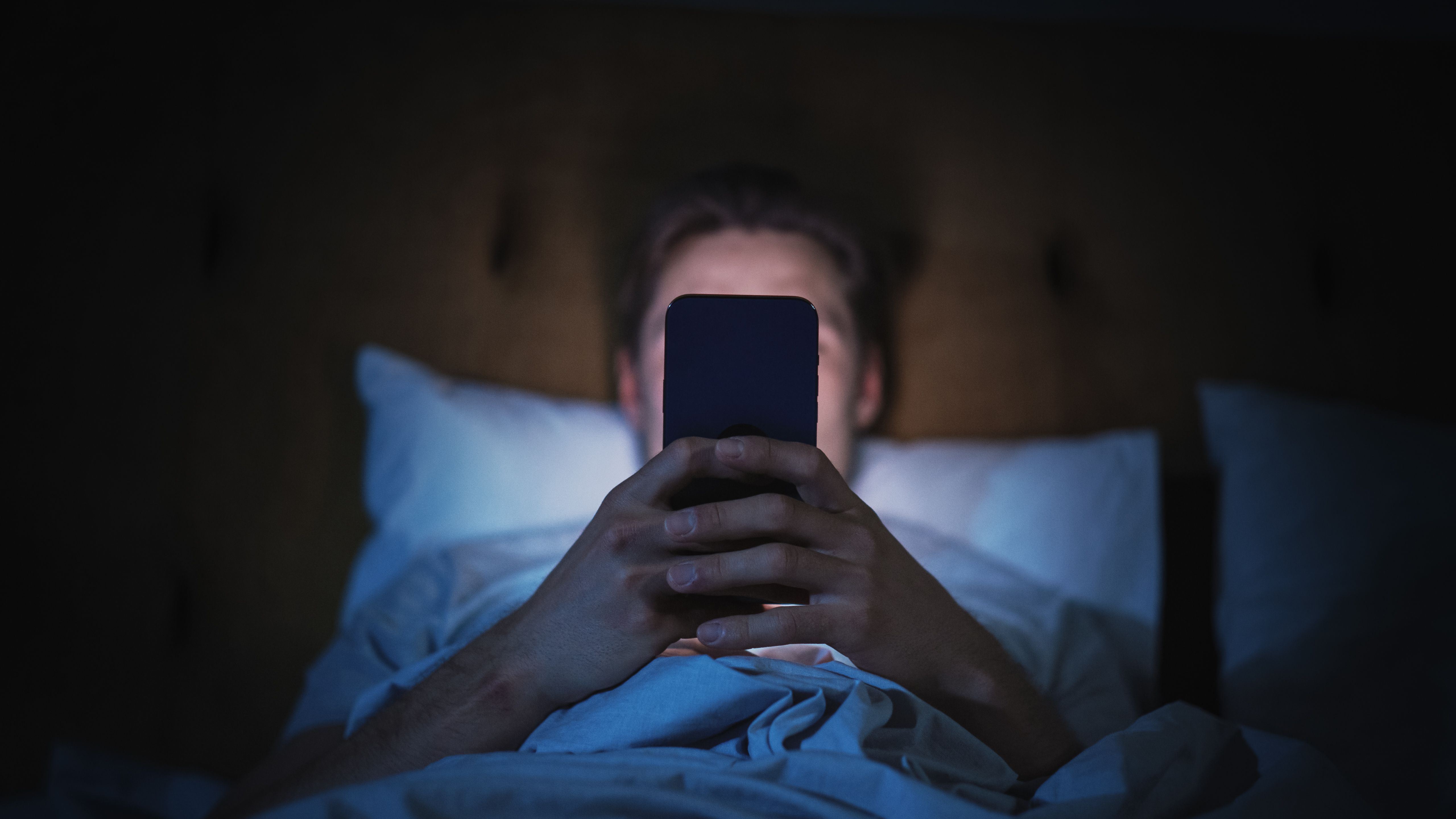 Anonymous Man Uses Smartphone in Bed at Home at Night. Handsome Guy Browsing Social Media, Reading News, Doing Online Shopping Late at Night. Focus on Hand Holding Mobile Phone Covering Face