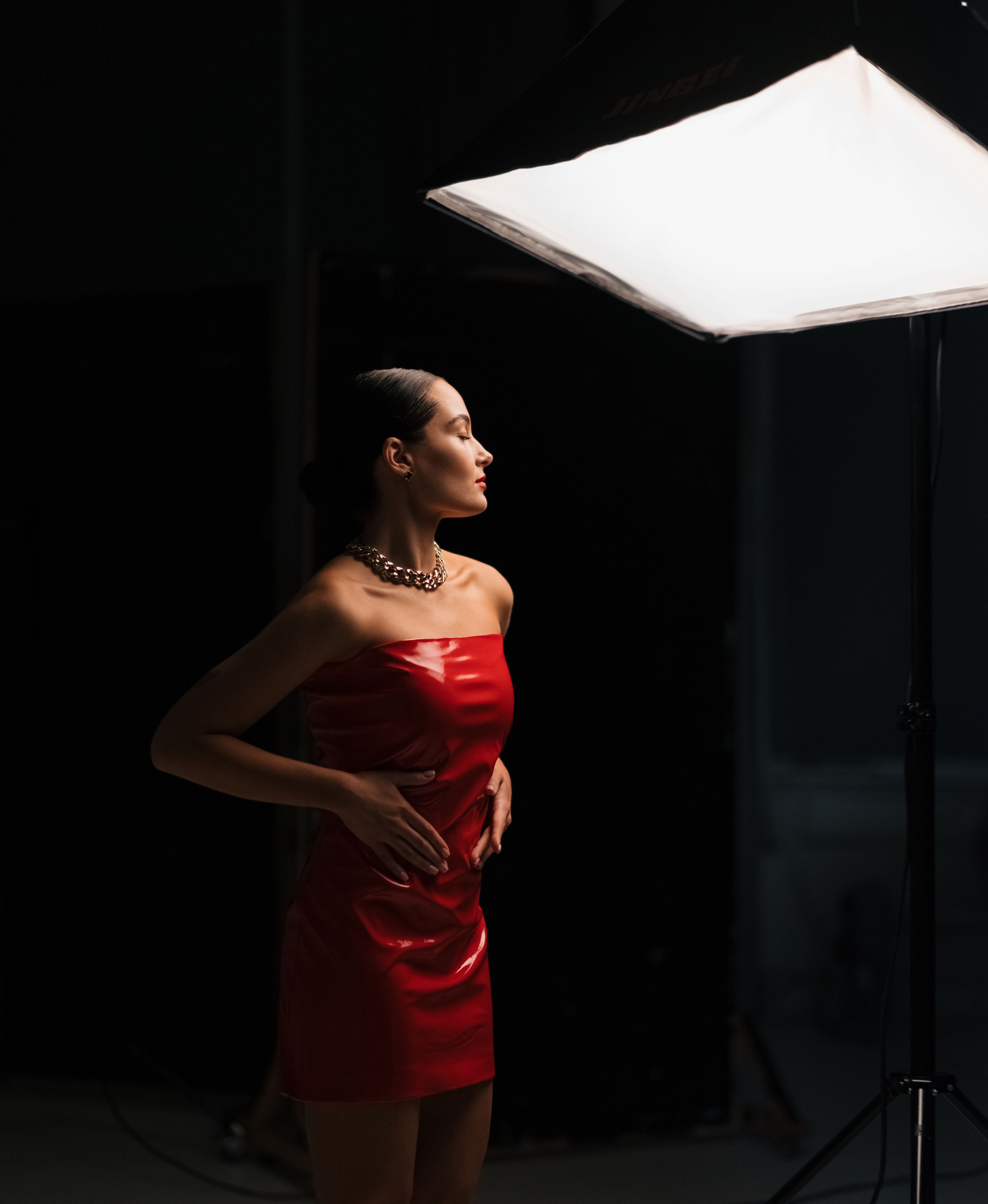 Young pretty woman poses in red latex dress under spotlight in studio.