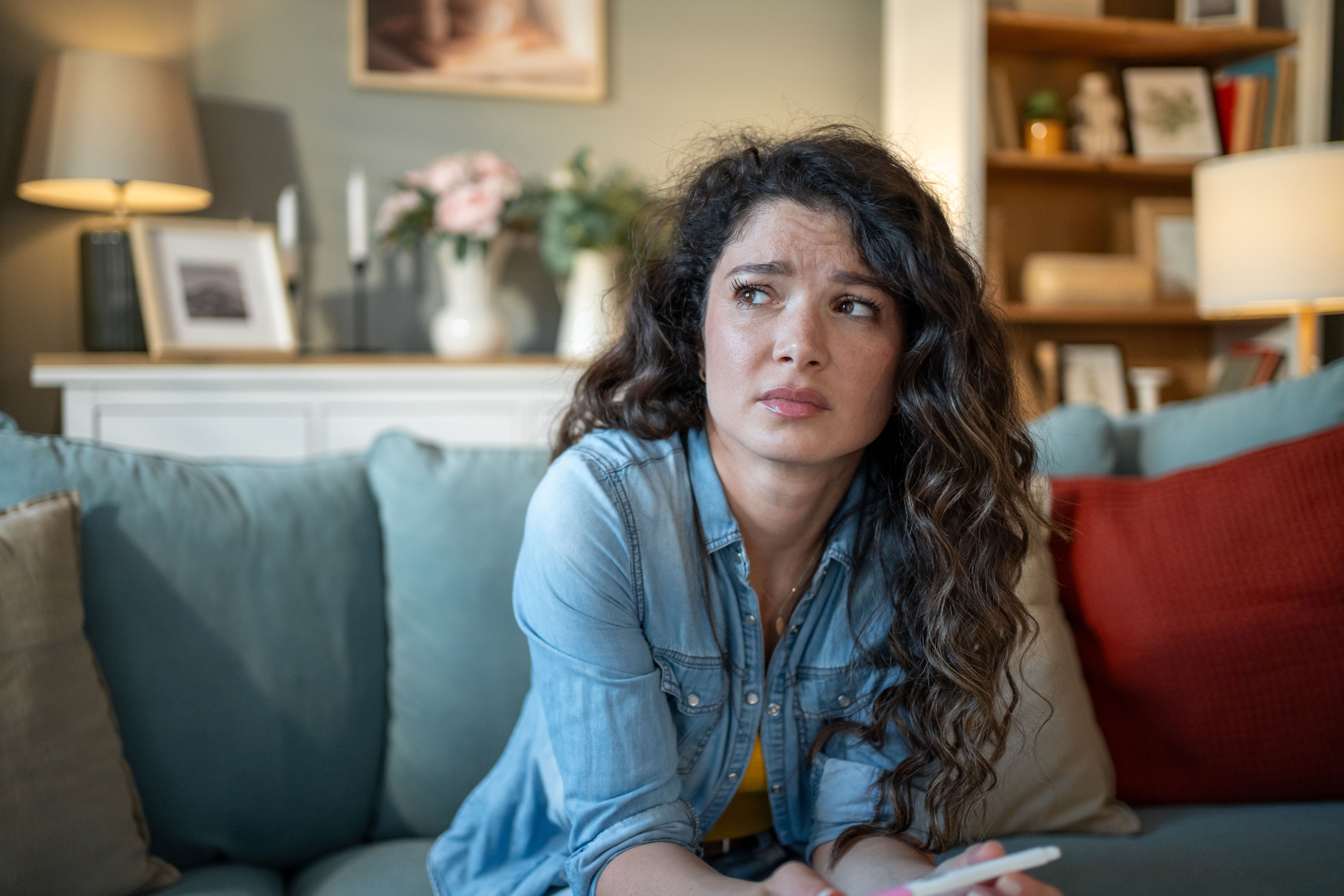 Worried young woman looking away while holding pregnancy test on sofa
