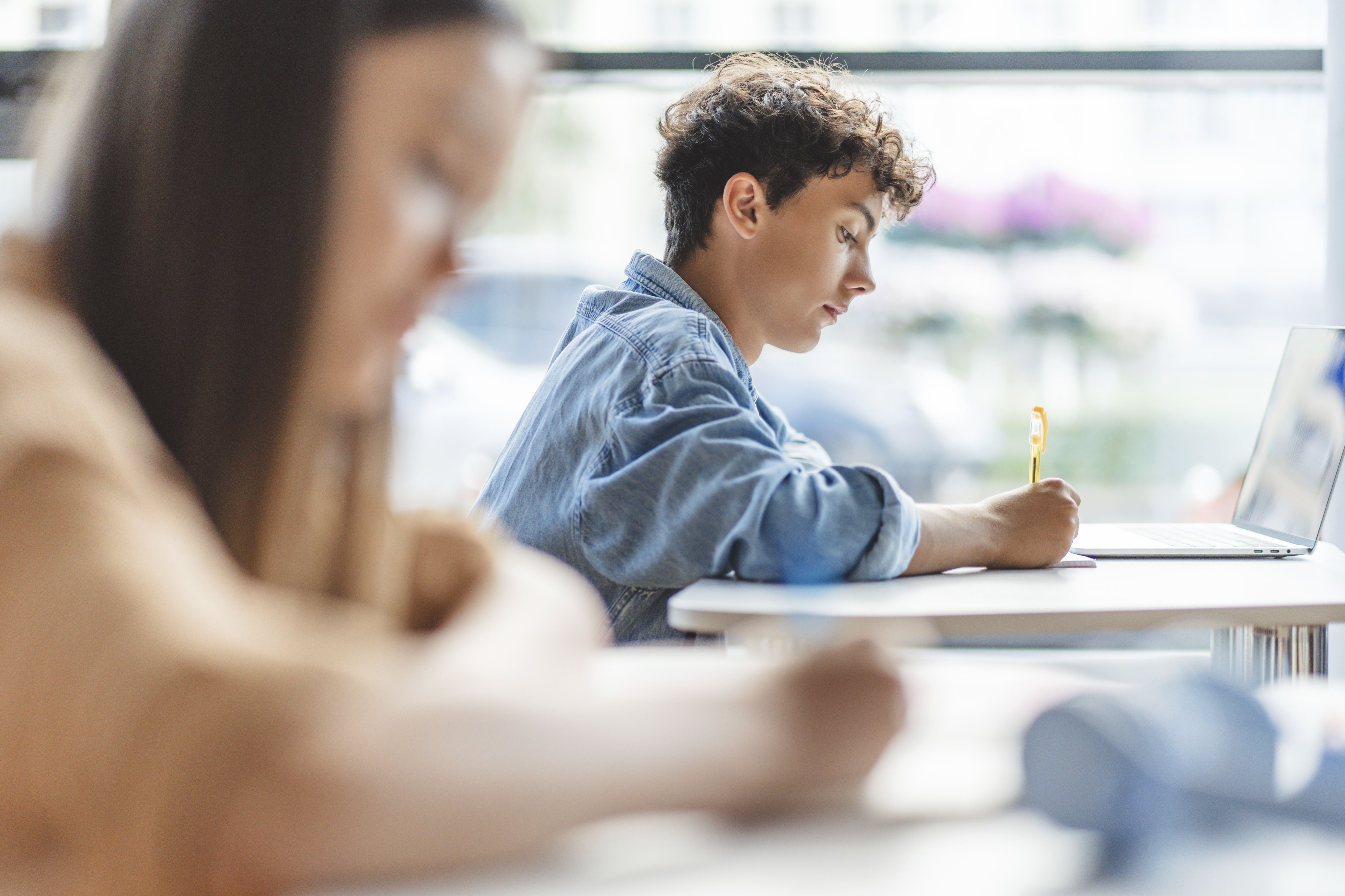 Pensive smart teenage boy using laptop taking notes in classroom. Back to school, education concept