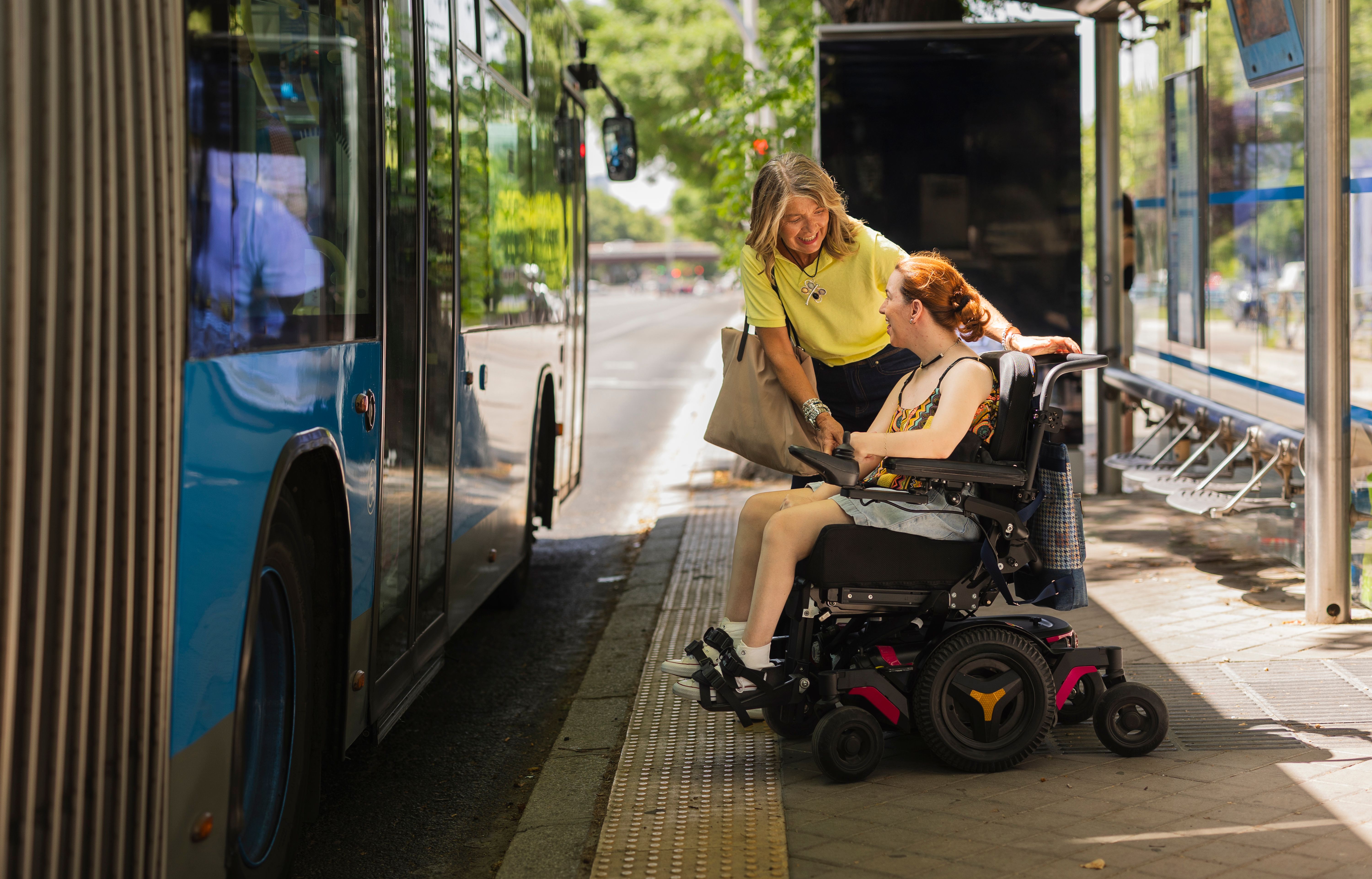 Woman helping disabled girl on wheelchair to board bus at bus stop