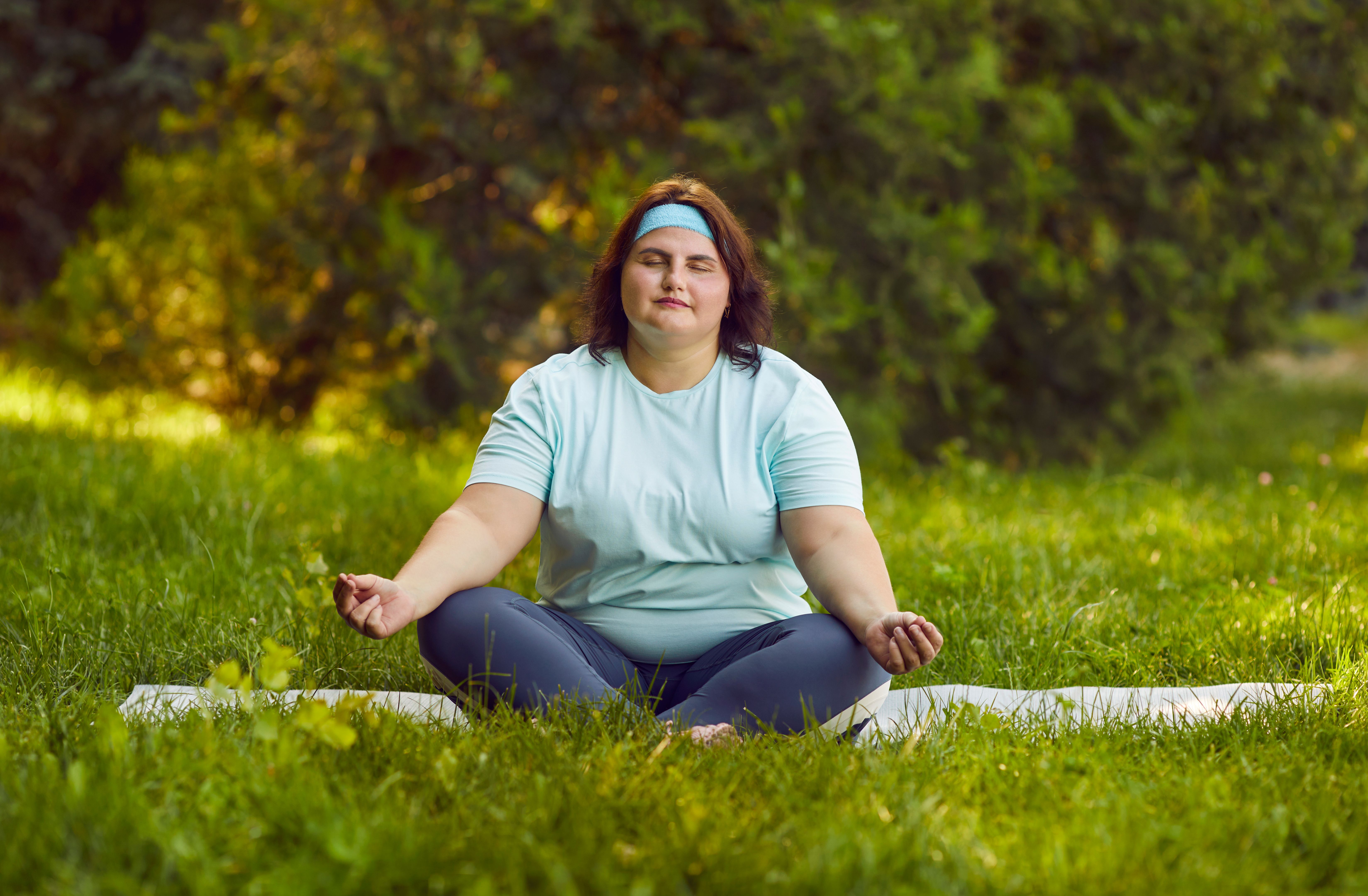 Overweight woman doing yoga in park, sitting on mat in grass, meditating and relaxing
