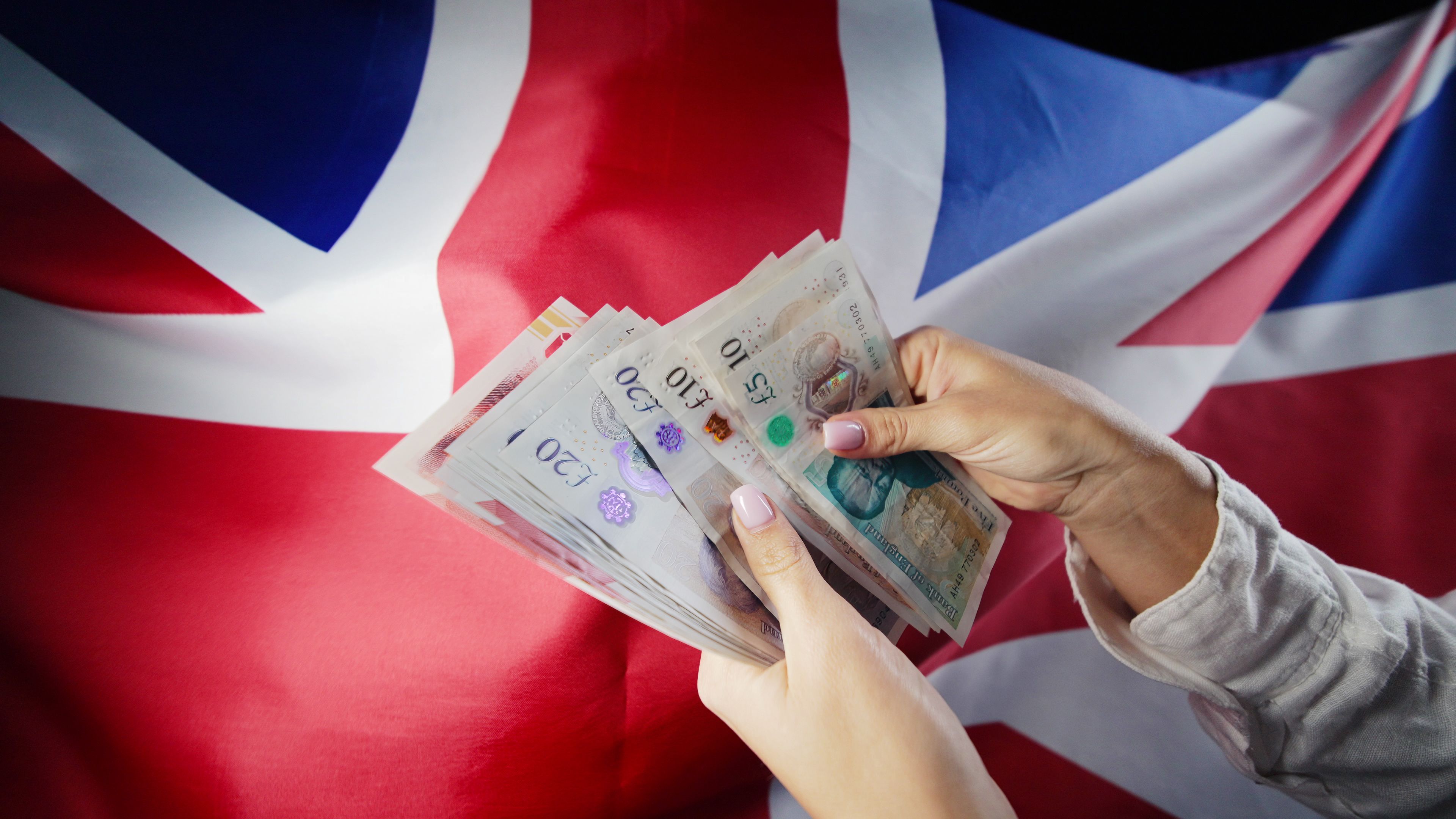 UK flag background. Woman counting GBP money pounds sterling. Credits, raising tariffs in Great Britain, traveling costs, financial concept. UK flag background. Woman counting GBP money pounds sterling. Credits, raising tariffs in Great Britain, traveling costs, financial concept.
