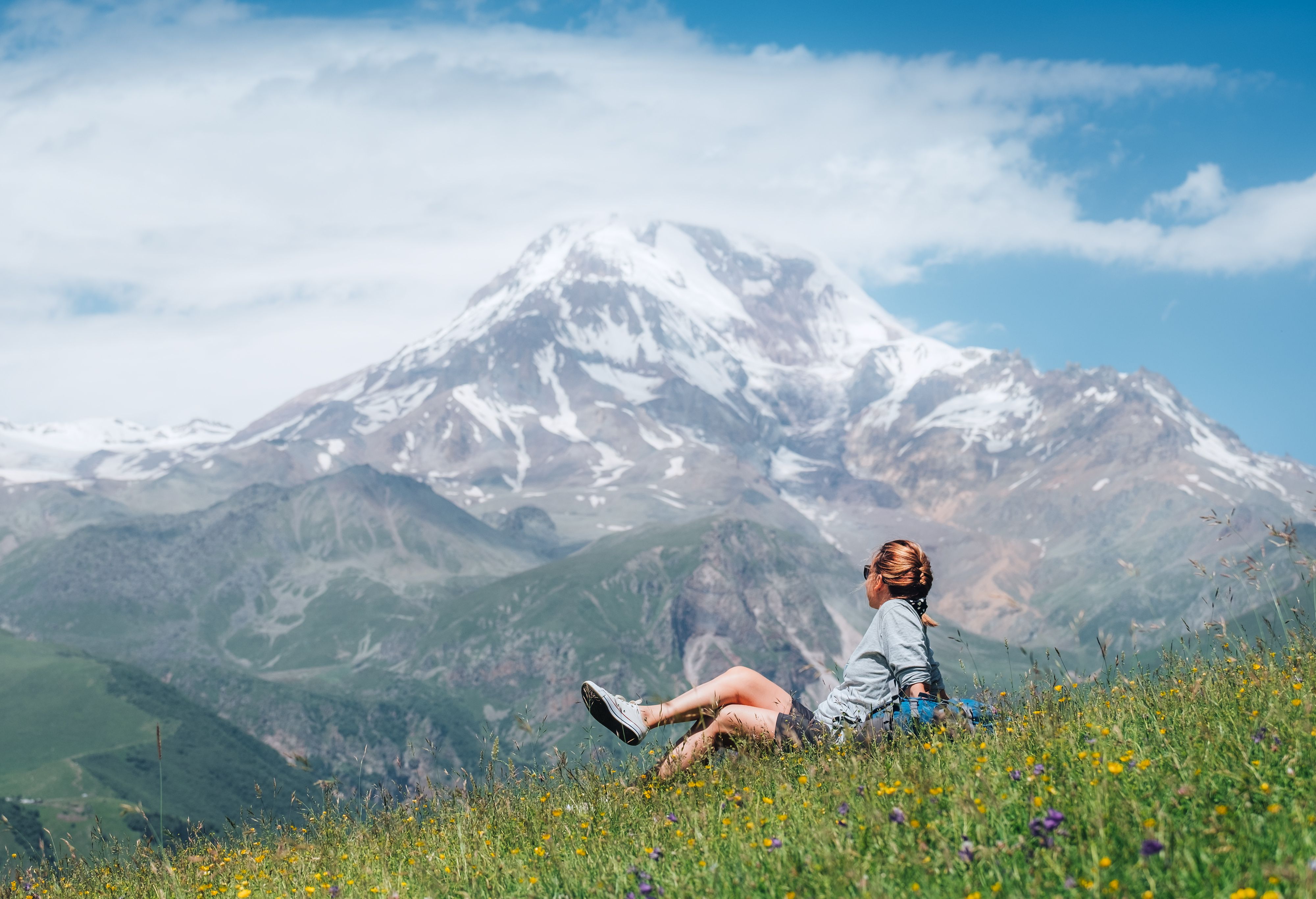Backpacker woman sitting on a green grass hill and enjoying snowy slopes of Kazbek 5054m mountain with a backpack while she walking by green grass hill. East Caucasus mountains, Georgia.