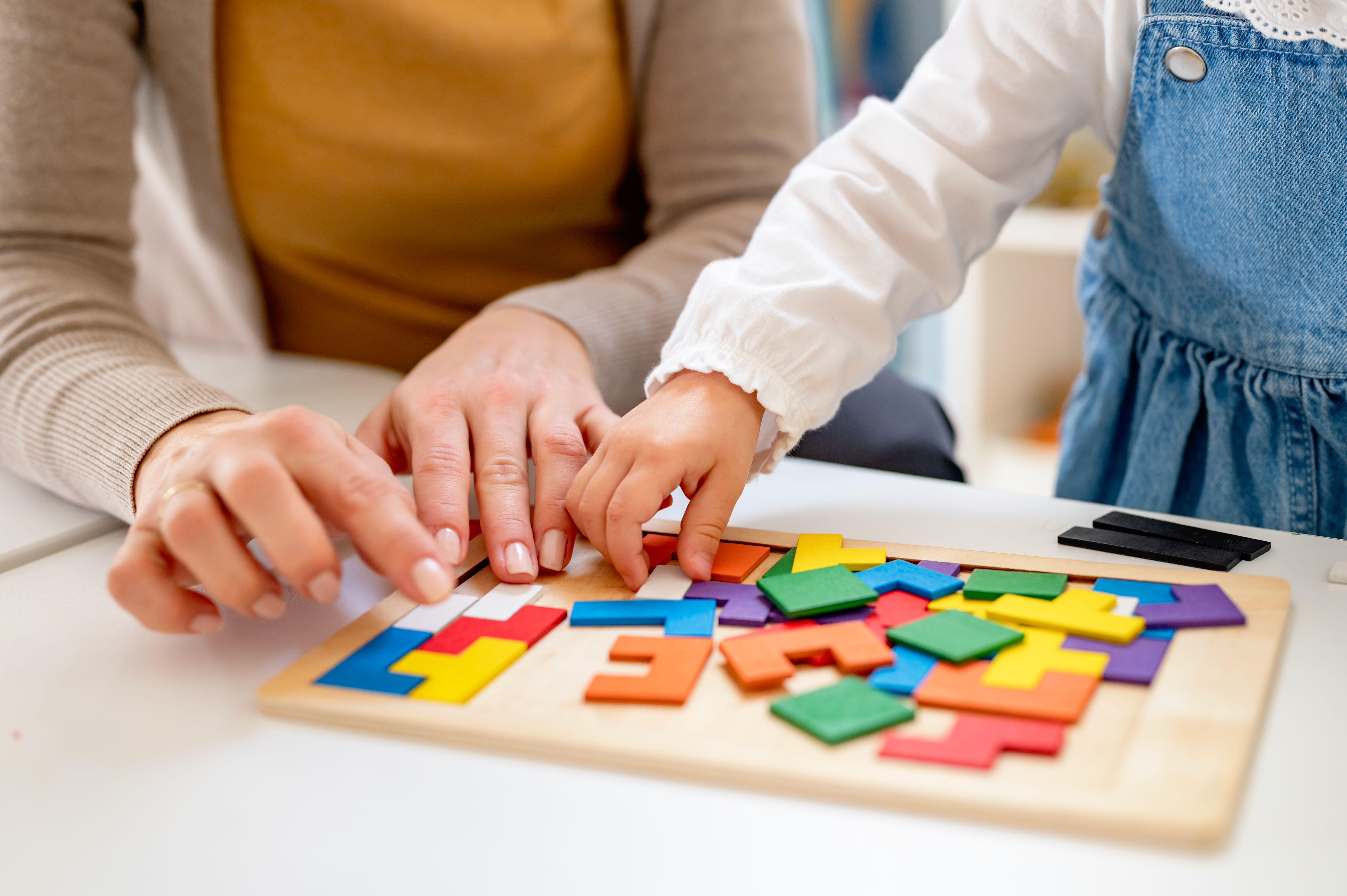 A caring preschool teacher helps a shy young girl with a colorful puzzle, encouraging her to adapt to the classroom environment and develop problem-solving skills through play-based learning