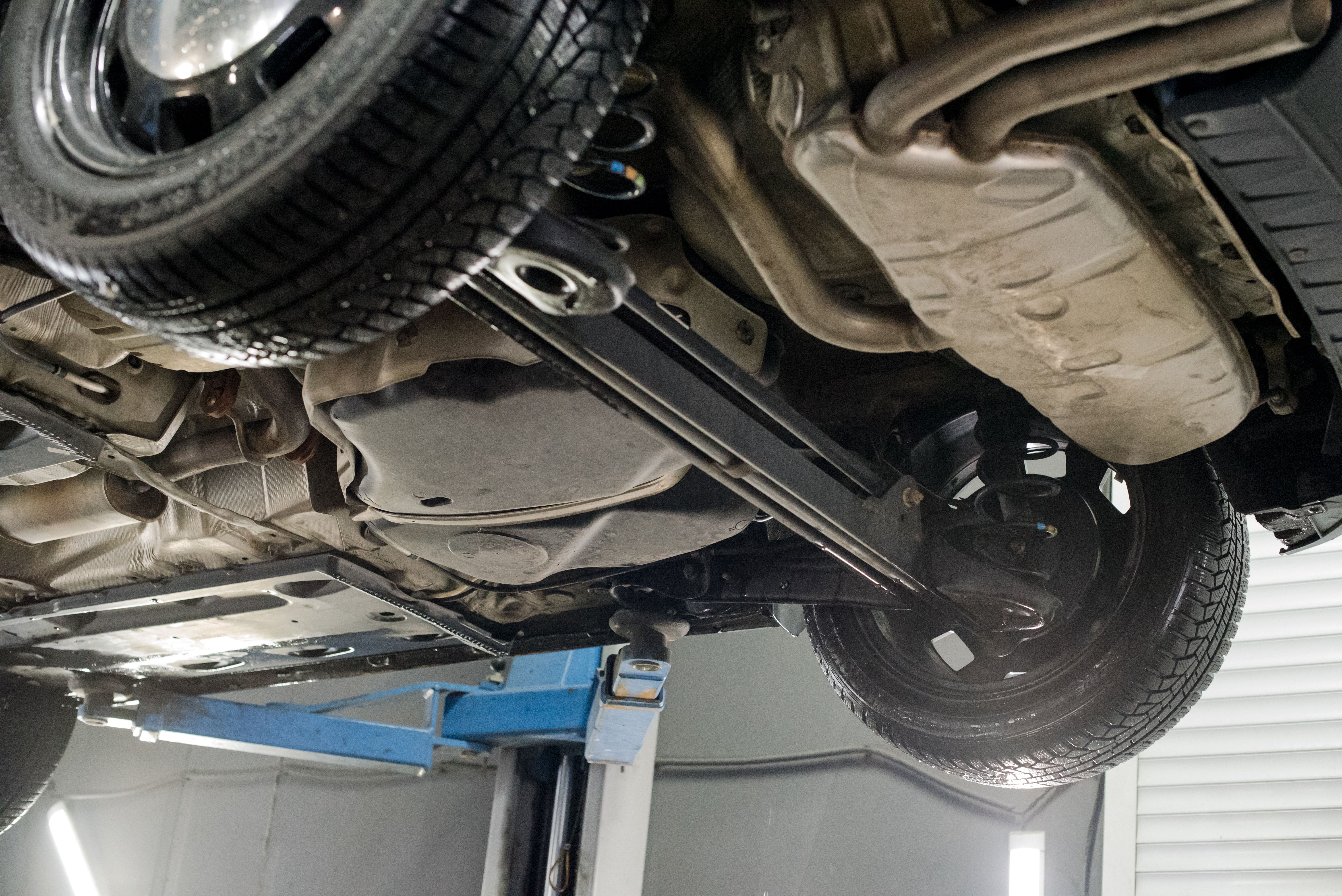 Modern car lifted on a hydraulic platform in an auto repair shop showing its undercarriage, exhaust system, and rear suspension while a technician performs maintenance work underneath.
