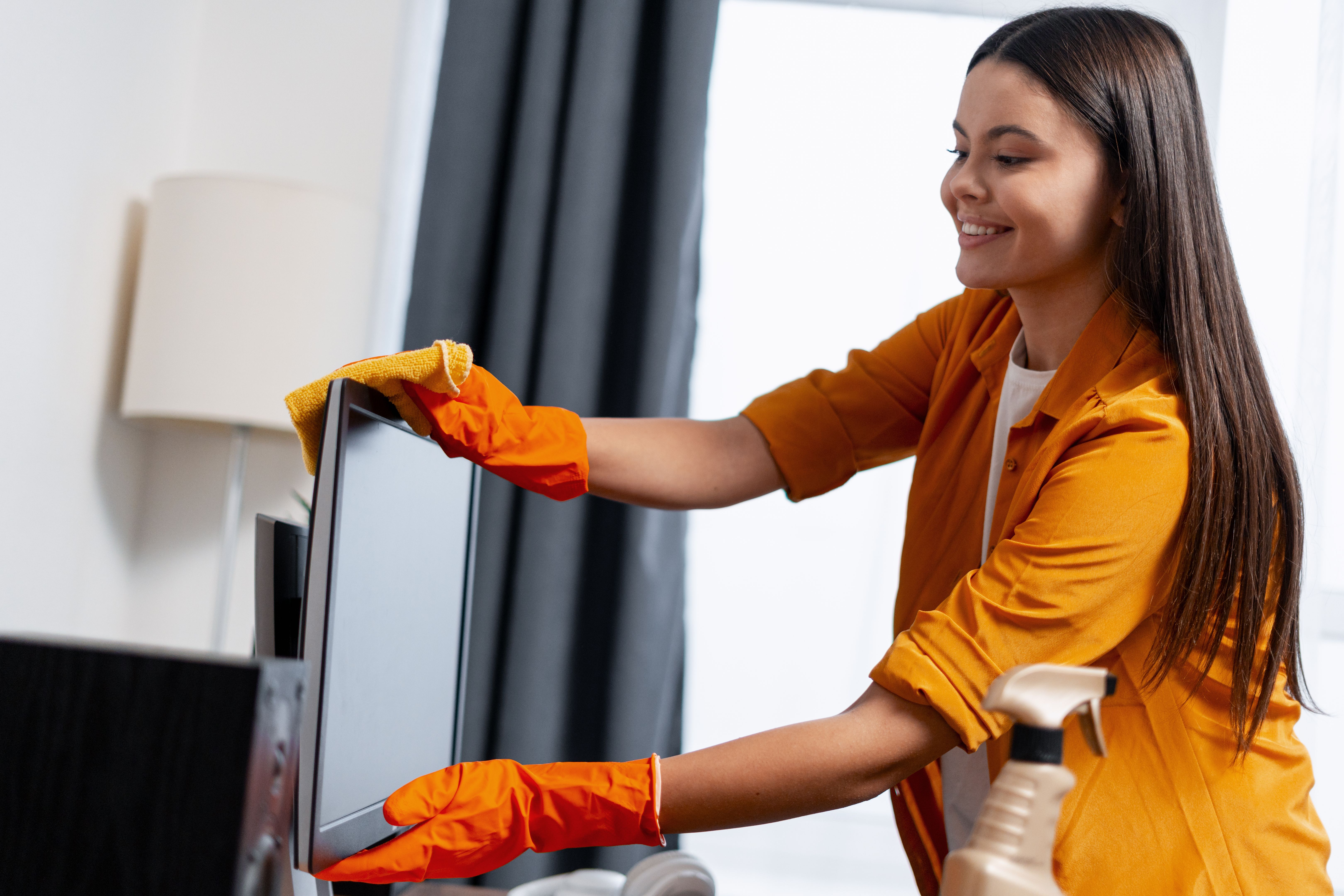 Young woman smiling while cleaning television screen at home, wearing orange gloves