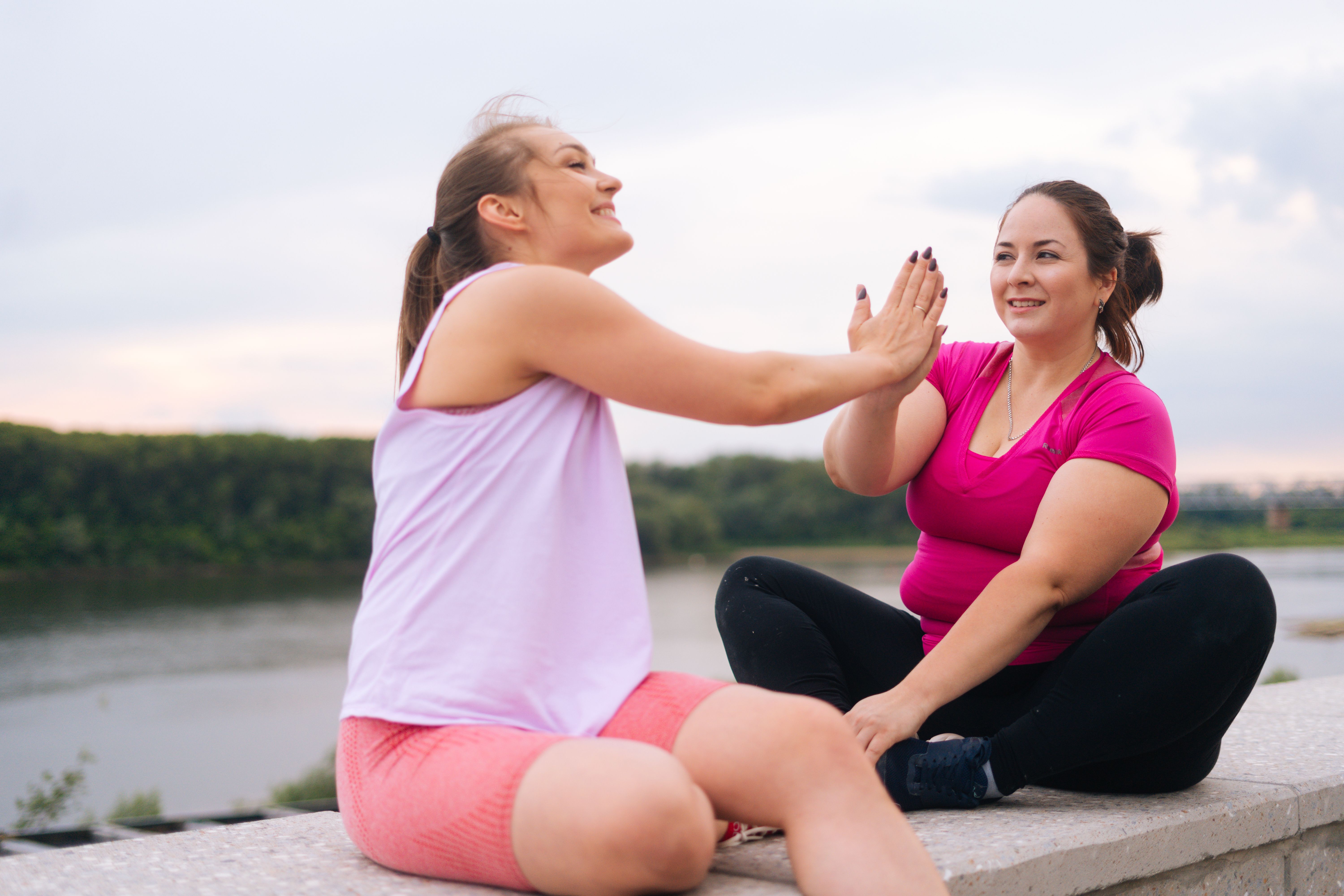 Cheerful slim fitness female trainer and motivated fat young woman greeting each other with a high-five other after exercising outdoors.