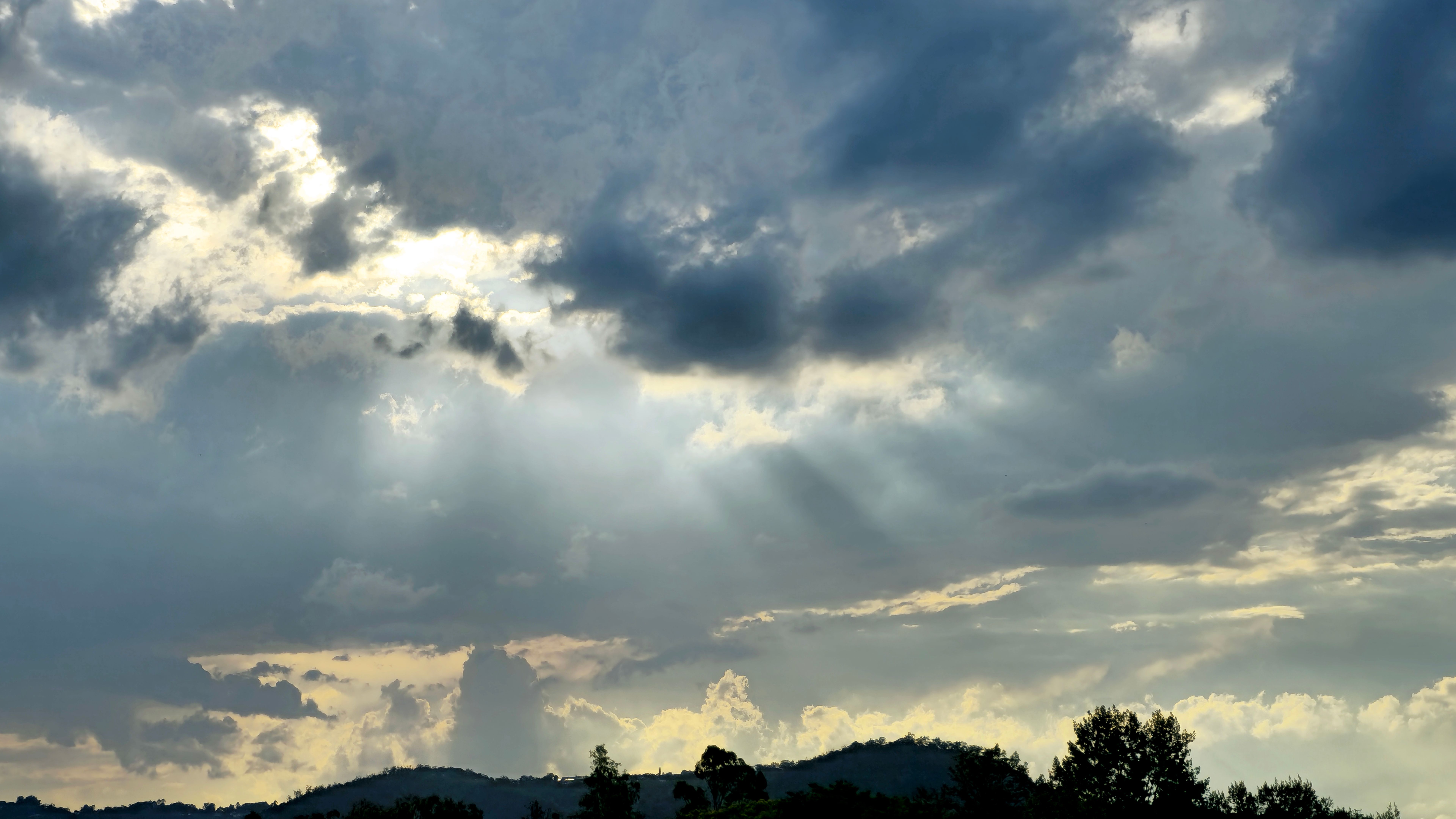 Cloudy sky with sun rays breaking through the clouds, illuminating the landscape below. The scene includes silhouetted trees and hills, creating a dramatic and serene atmosphere. Cloudy sky with sun rays breaking through the clouds, illuminating the landscape below. The scene includes silhouetted trees and hills, creating a dramatic and serene atmosphere.