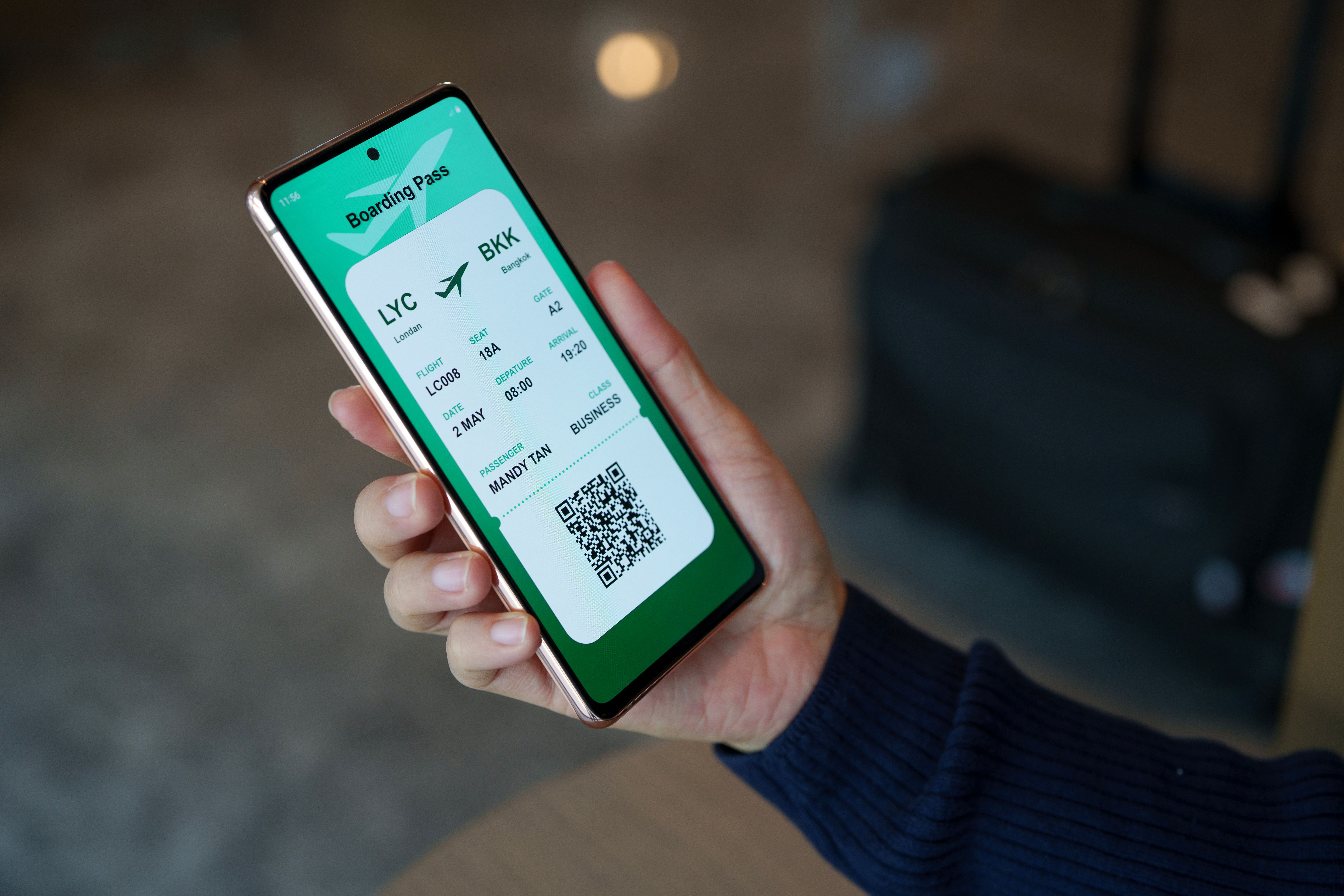 A close-up image shows a woman sitting in the waiting lounge, holding a smartphone displaying an electronic flight ticket as she completes an online check-in for her vacation A close-up image shows a woman sitting in the waiting lounge, holding a smartphone displaying an electronic flight ticket as she completes an online check-in for her vacation