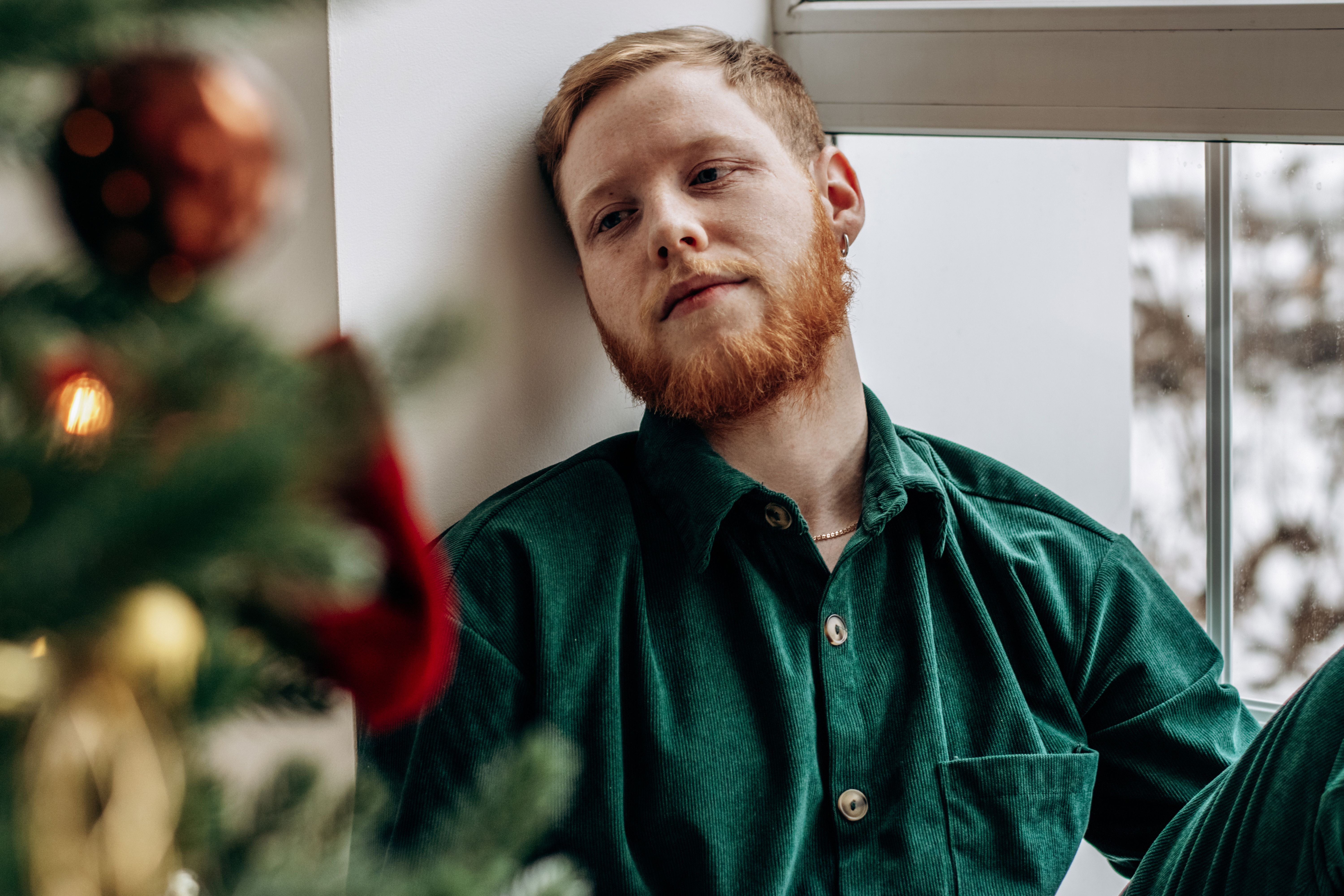 Attractive red-haired bearded young man is sitting by the window by the Christmas tree.New Year and Christmas concept Attractive red-haired bearded young man is sitting by the window by the Christmas tree.New Year and Christmas concept